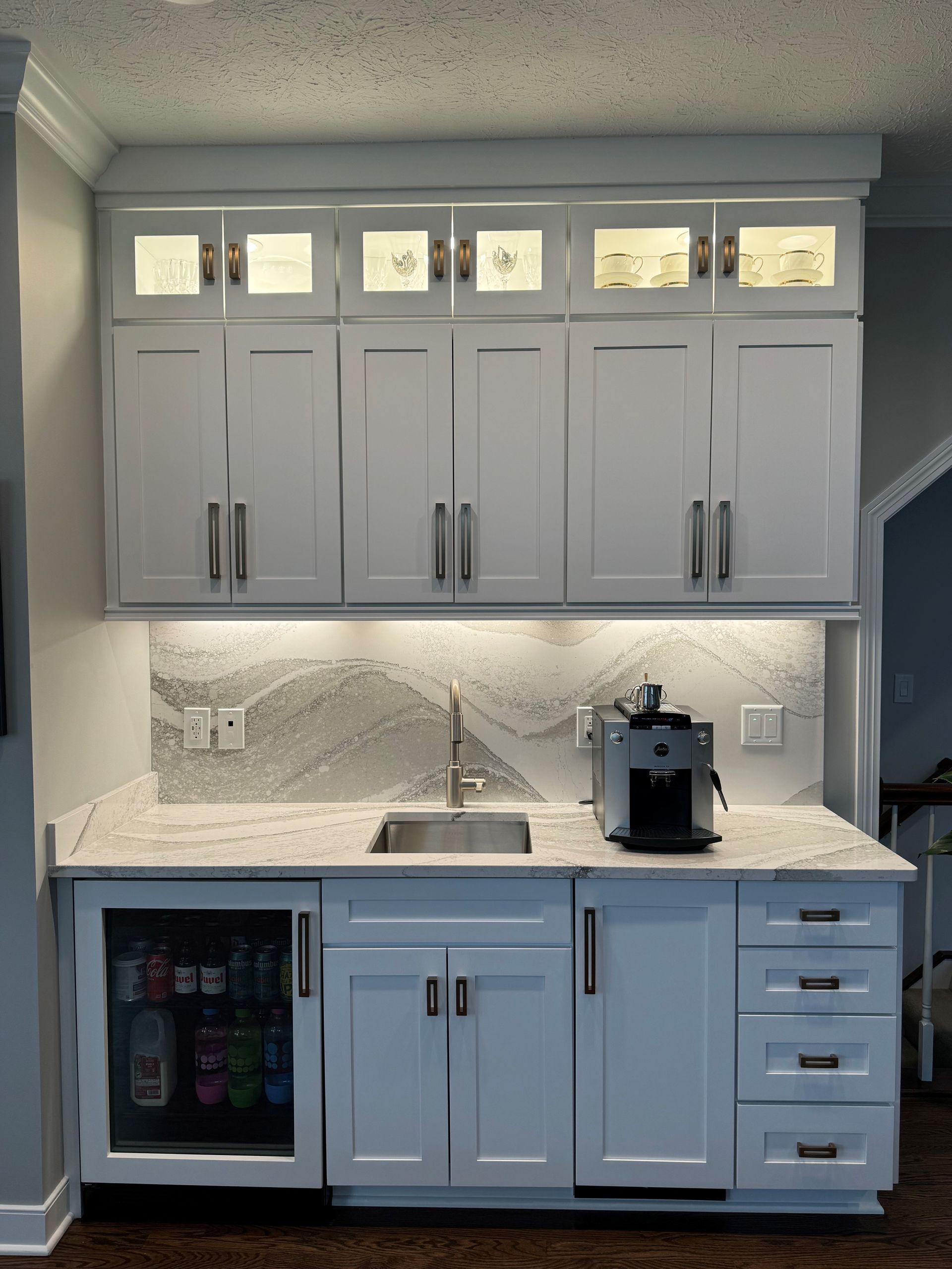 White built-in kitchen bar with upper cabinets, countertop, sink, mini-fridge, and coffee machine.