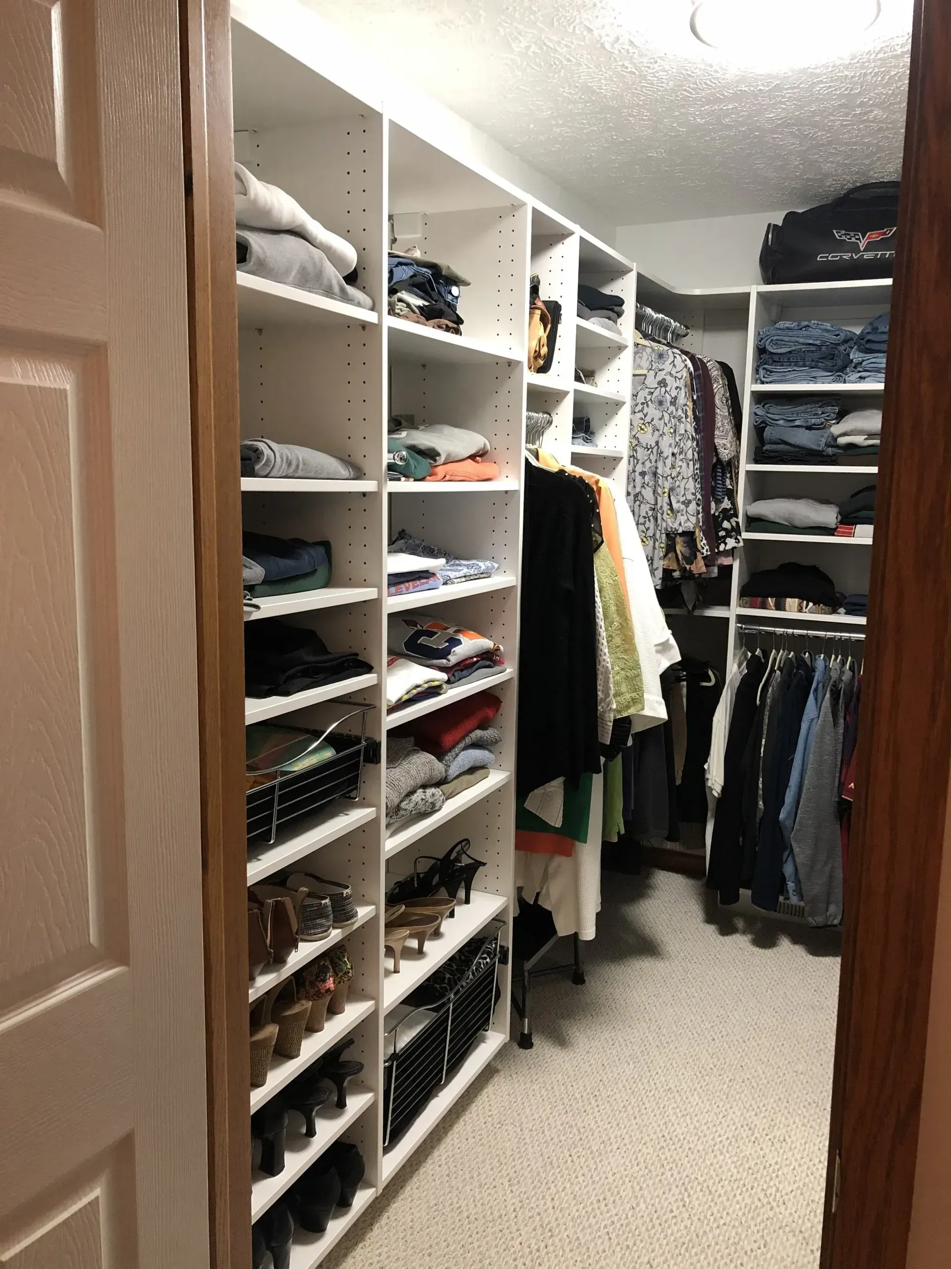 Walk-in closet with white shelves, clothing, shoes, and neatly folded items. Beige carpet, bright overhead lighting.