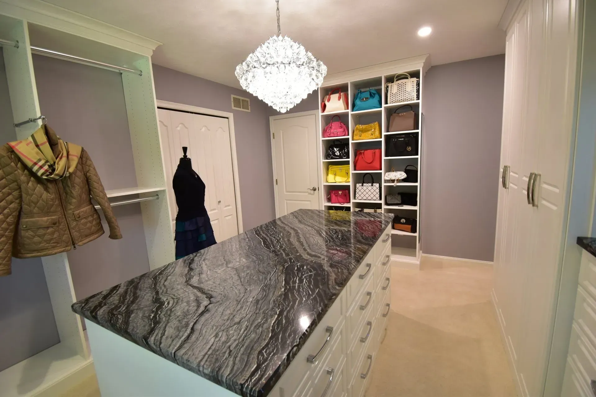 Walk-in closet with island, shelves of handbags, clothing rack with jacket, and crystal chandelier.