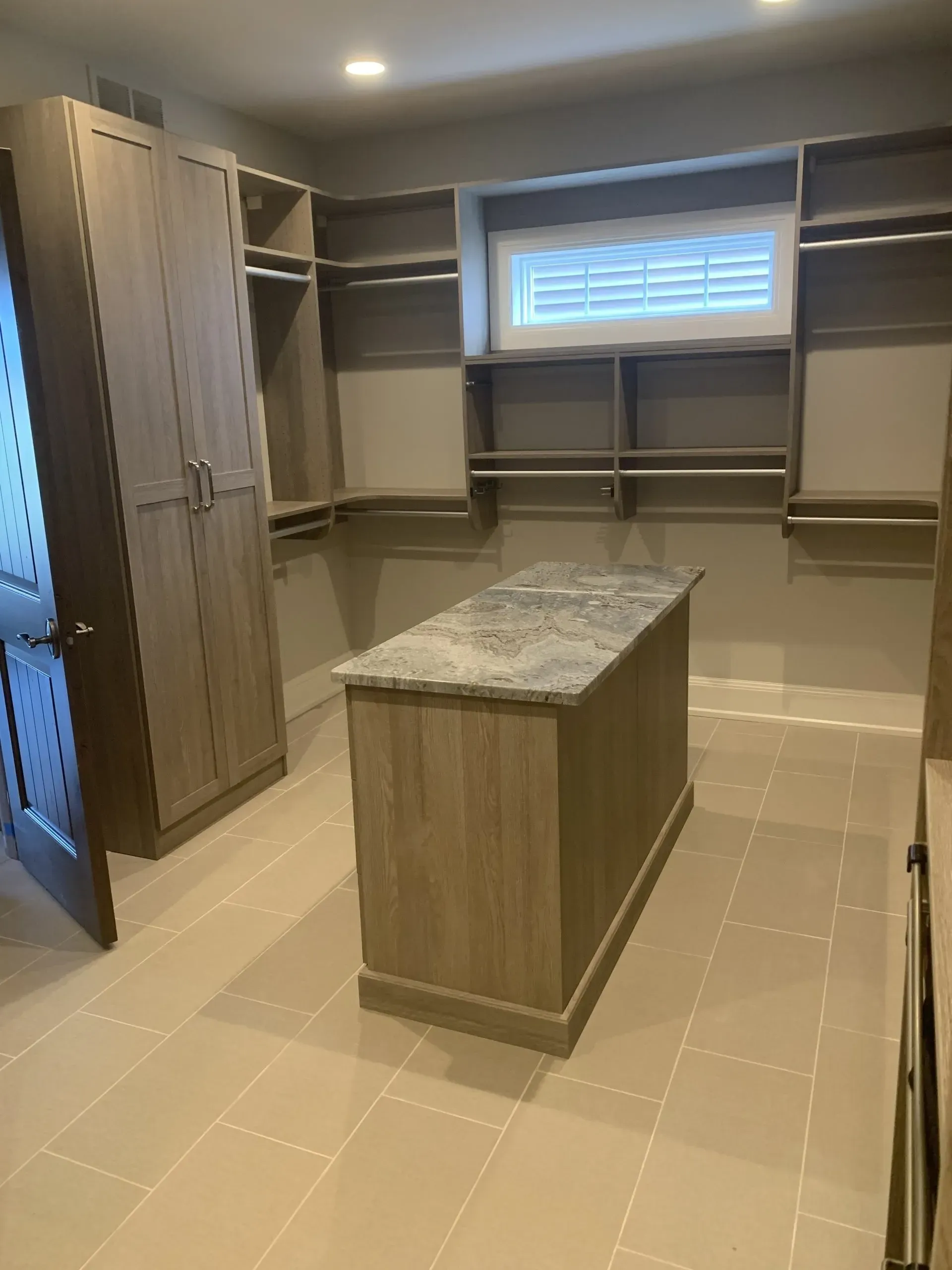 A walk-in closet with wooden cabinets and shelving, a center island with a marble top, and a small window.