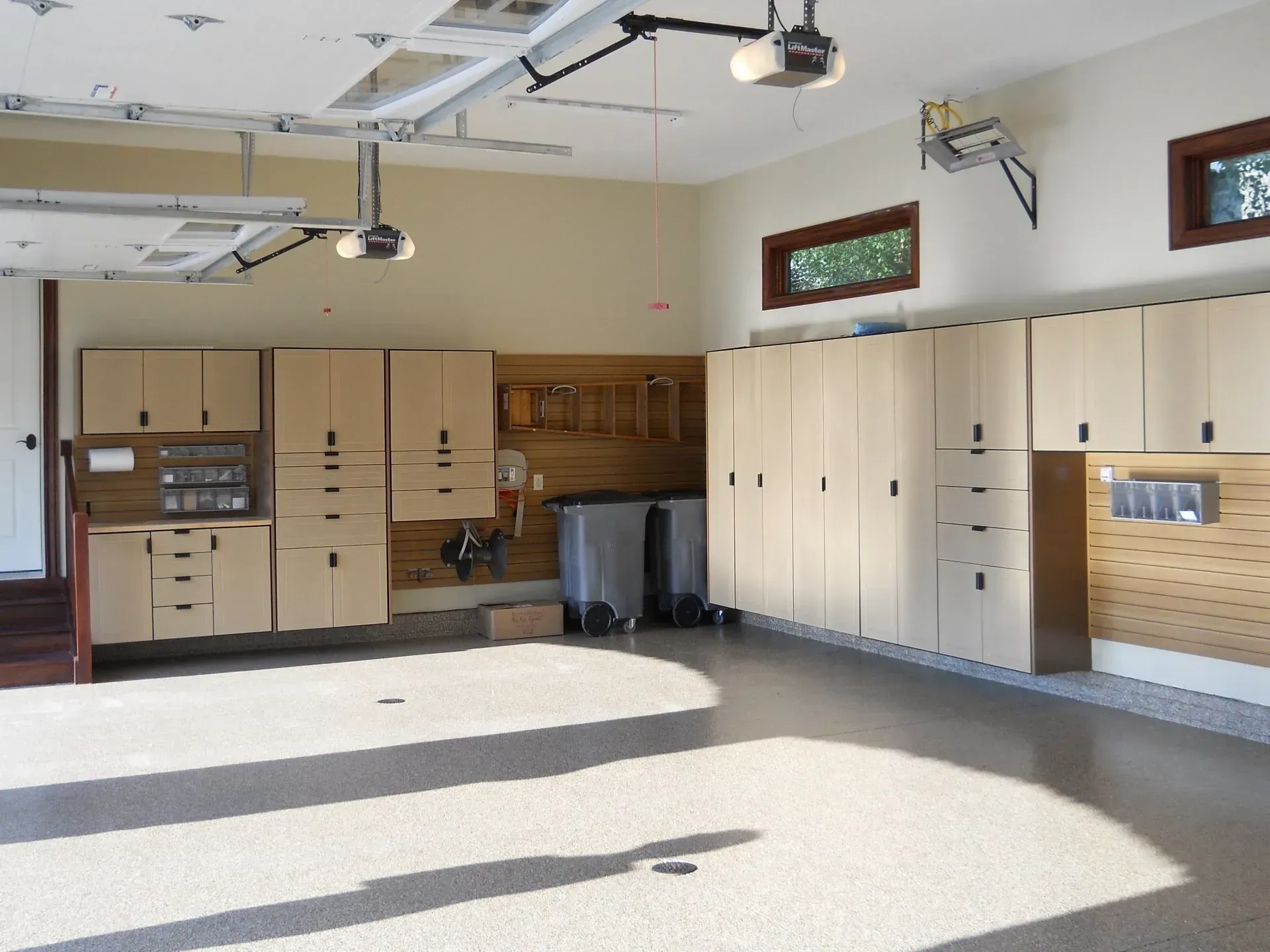 Spacious garage with light tan cabinets, a speckled floor, and two overhead garage door openers.