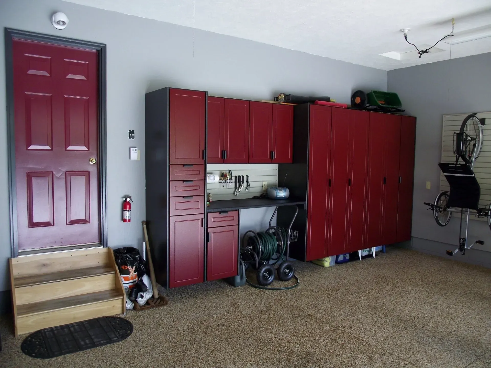 Red storage cabinets and door in a garage with gray walls, tan flooring, and work area.