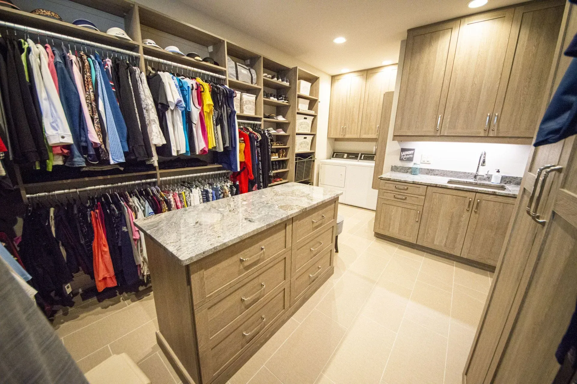 Walk-in closet with clothes, island drawers, and laundry area. Beige cabinets, light fixtures.