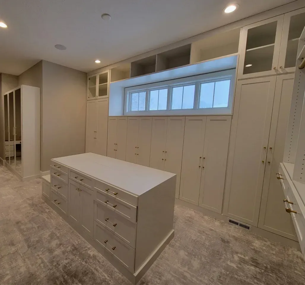 Walk-in closet with white cabinets, drawers, and a center island. A window is above the cabinets.