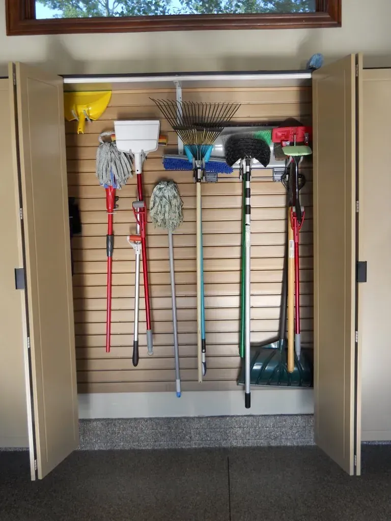 Garage closet with cleaning tools hung on a slotted wall; tan doors, various mop and broom colors.