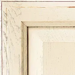Corner of a cream-colored, distressed wooden cabinet door with a raised frame.