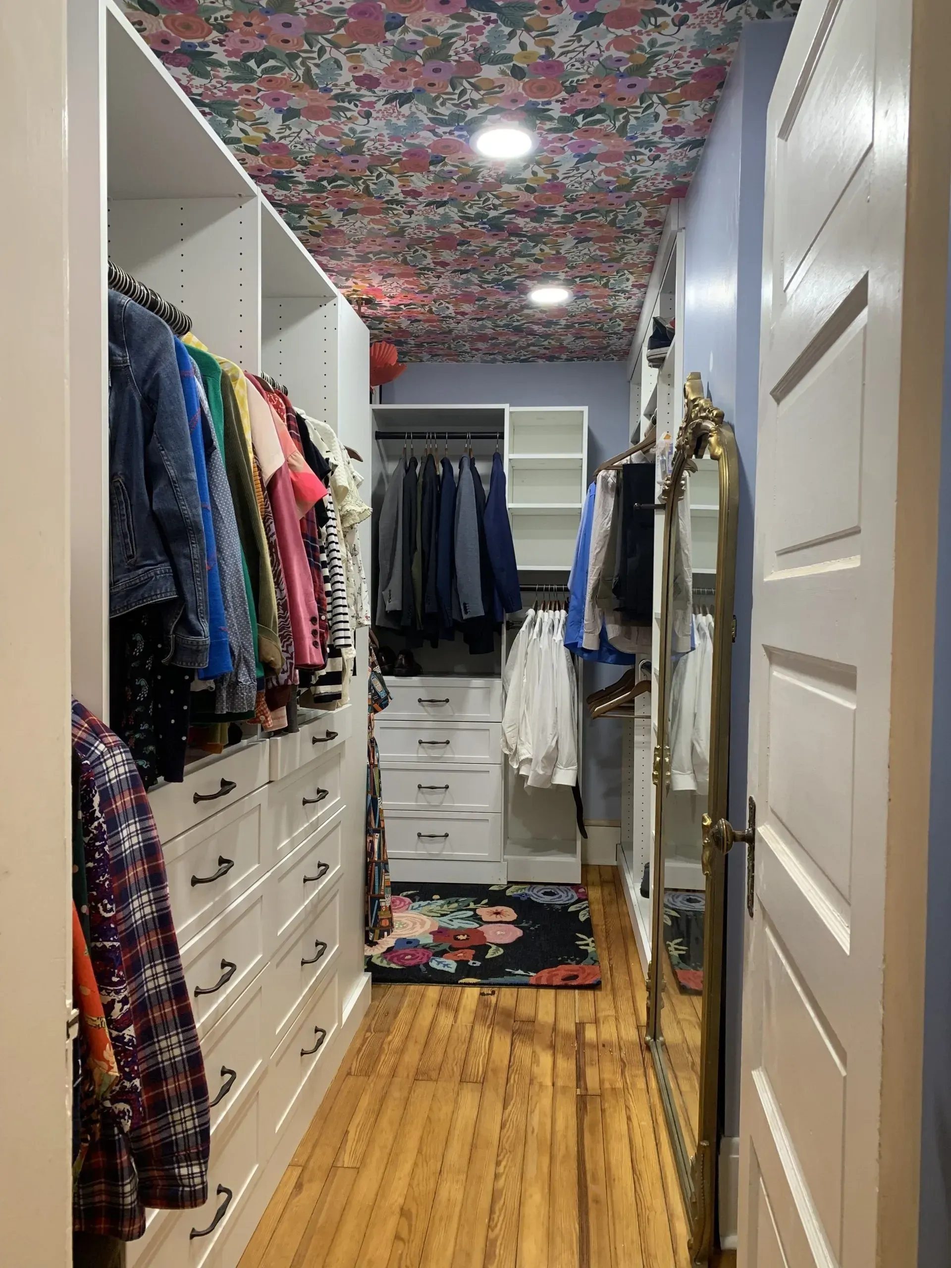 Narrow walk-in closet with white drawers, clothes hanging on rods, floral ceiling, and tall mirror.