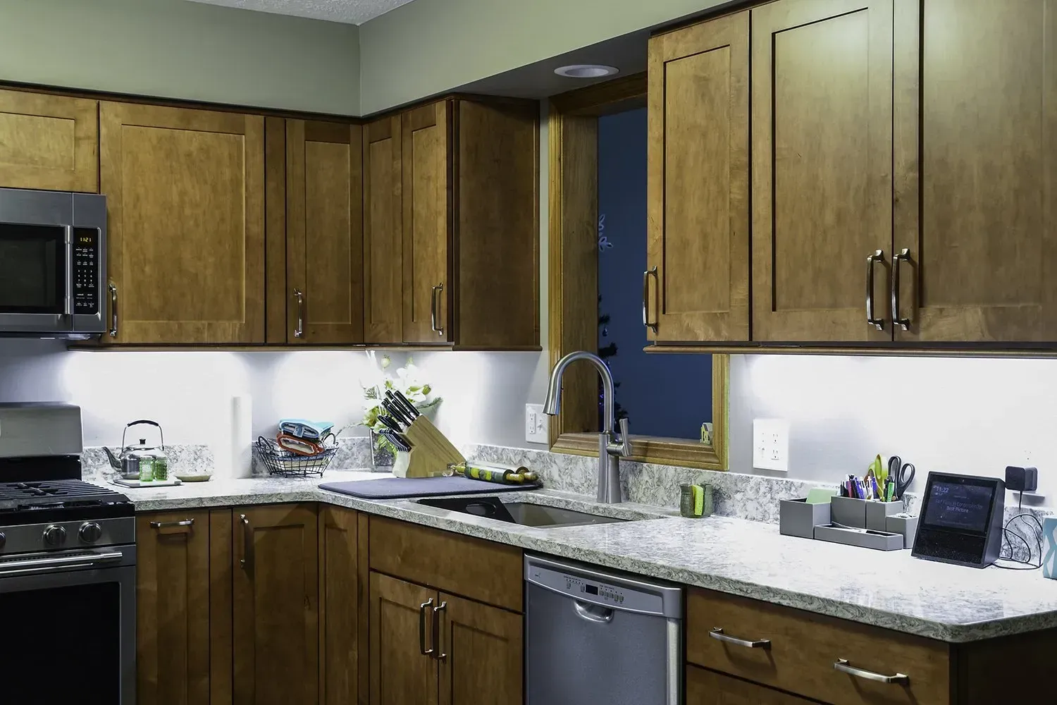 Wooden kitchen cabinets with under-cabinet lighting over a granite countertop, stainless steel appliances, and a window.