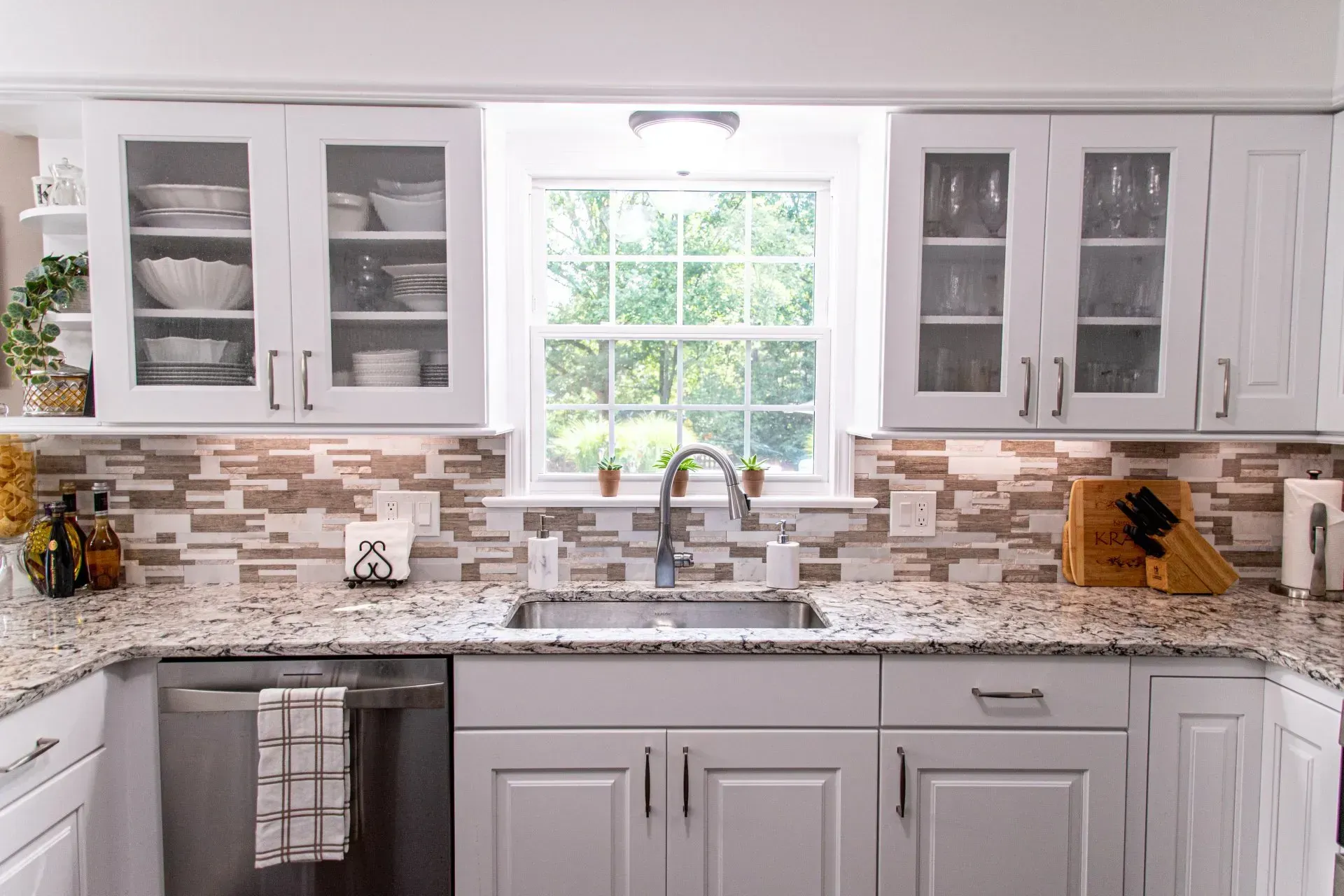 White kitchen with a window, stainless steel sink, and white cabinets with glass doors.