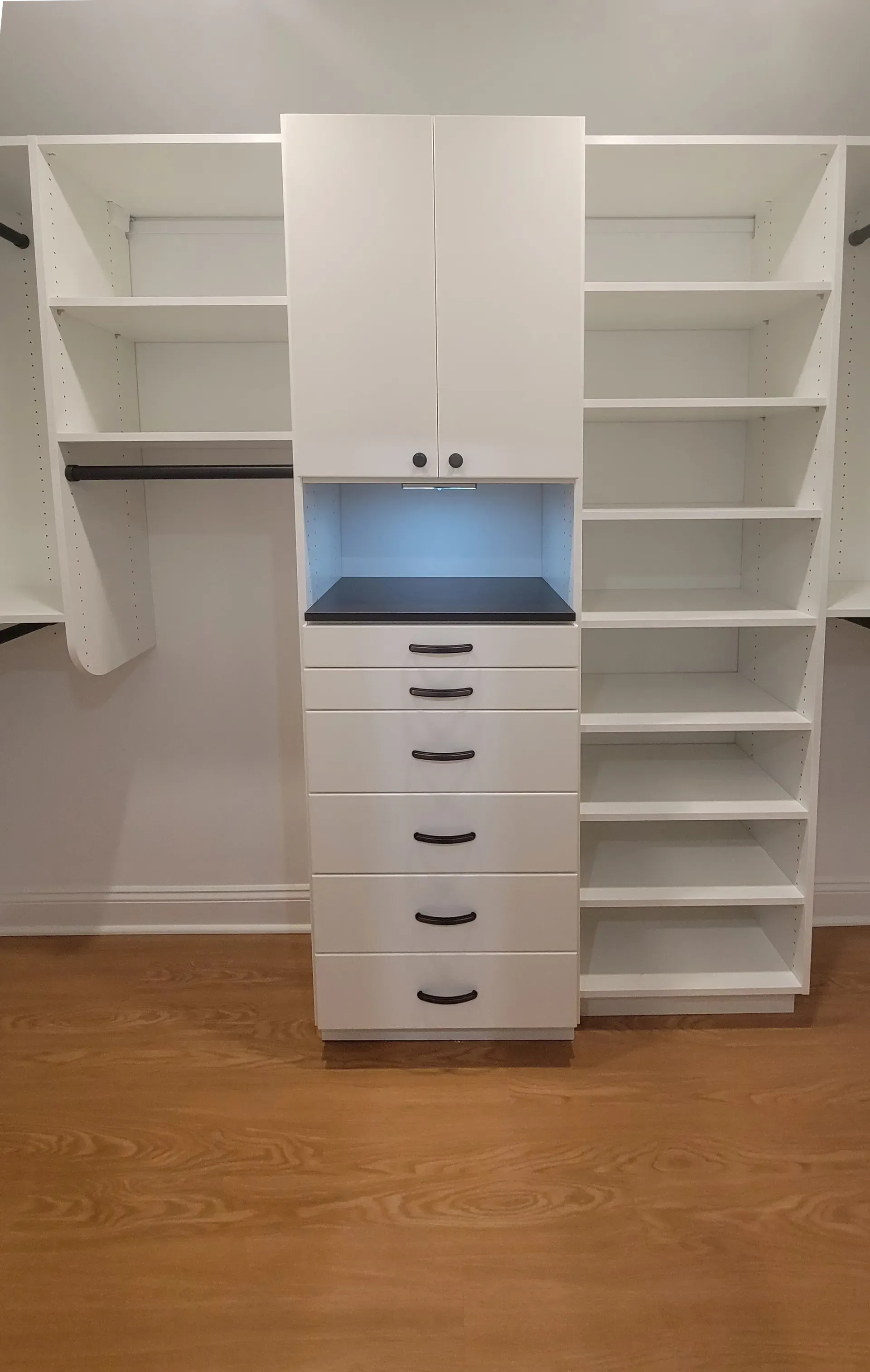 White closet organizer with shelves, drawers, and hanging rod, set against a wooden floor.