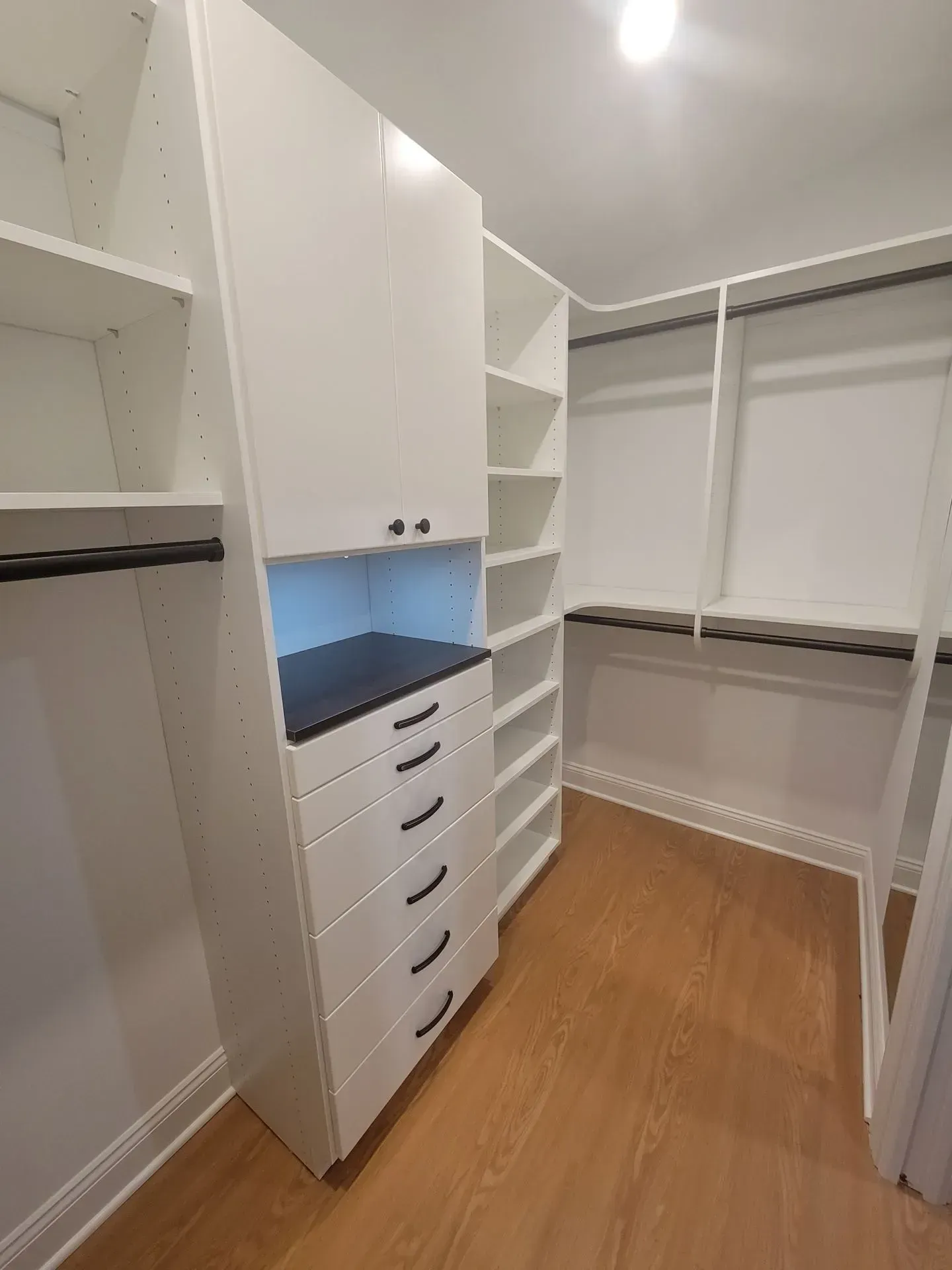 White walk-in closet with shelves, drawers, and hanging rods. Wooden floor, overhead lighting.