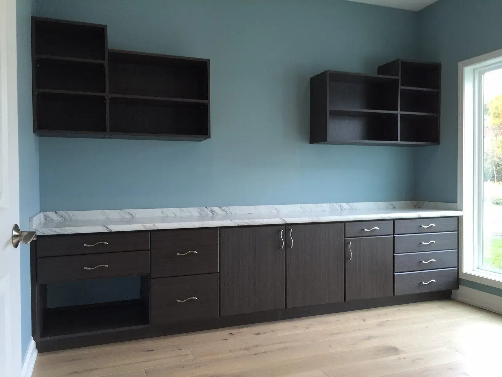 Dark cabinets and shelves on a blue wall with a white countertop and a window.
