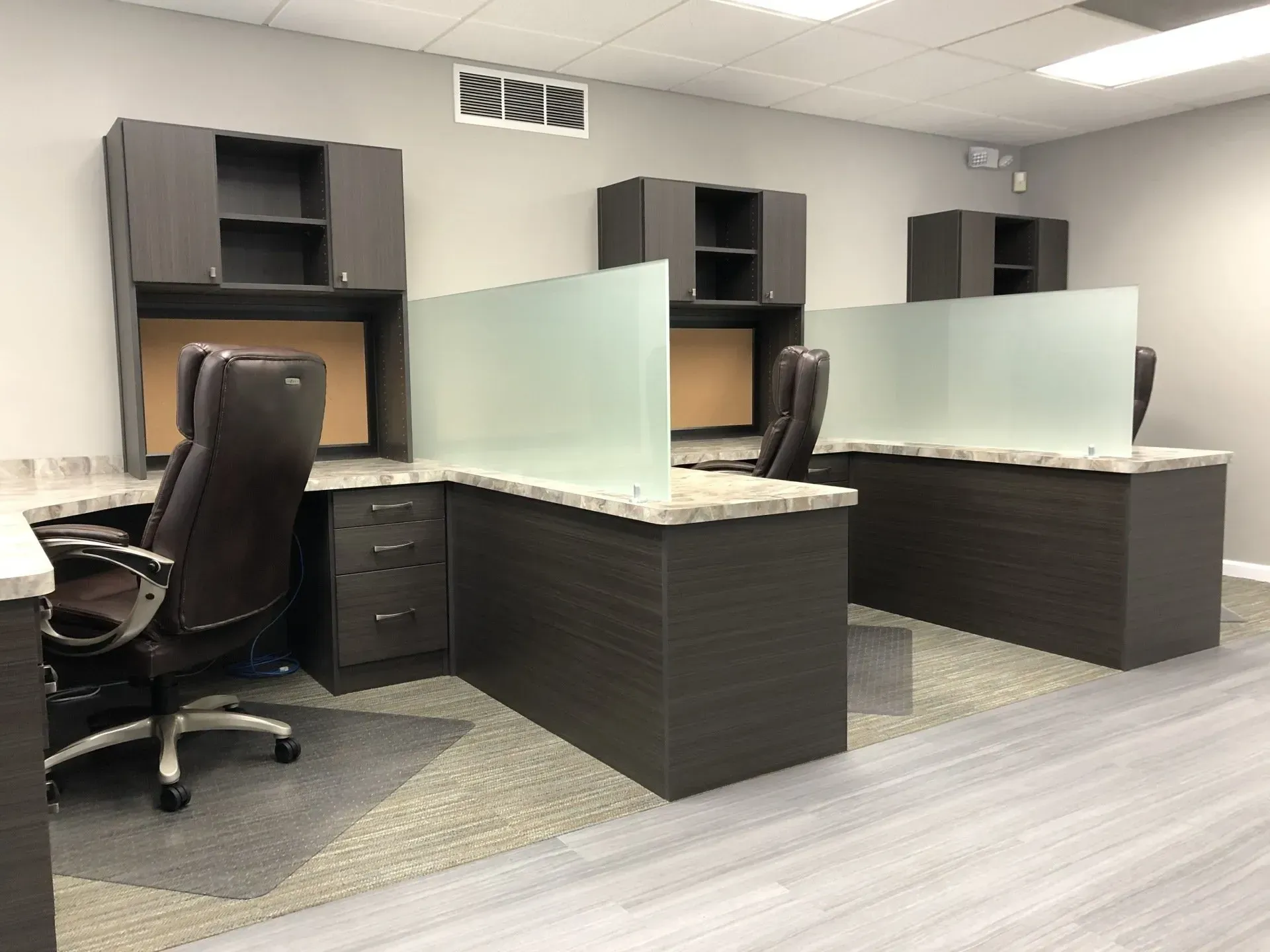 Dark gray office desks with frosted glass dividers and overhead cabinets.