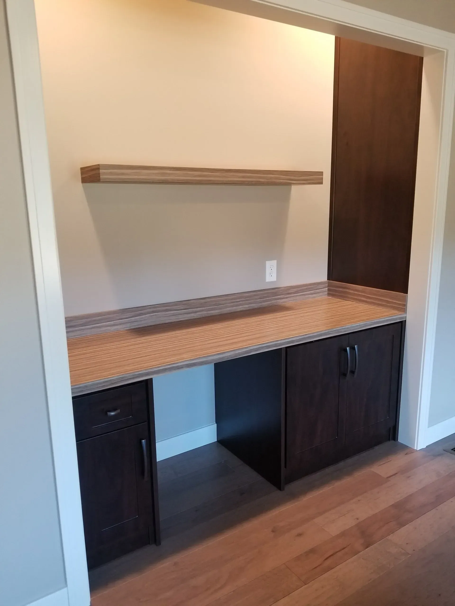Built-in home office: dark brown desk with countertop, cabinets, shelf; tan walls, hardwood floors.
