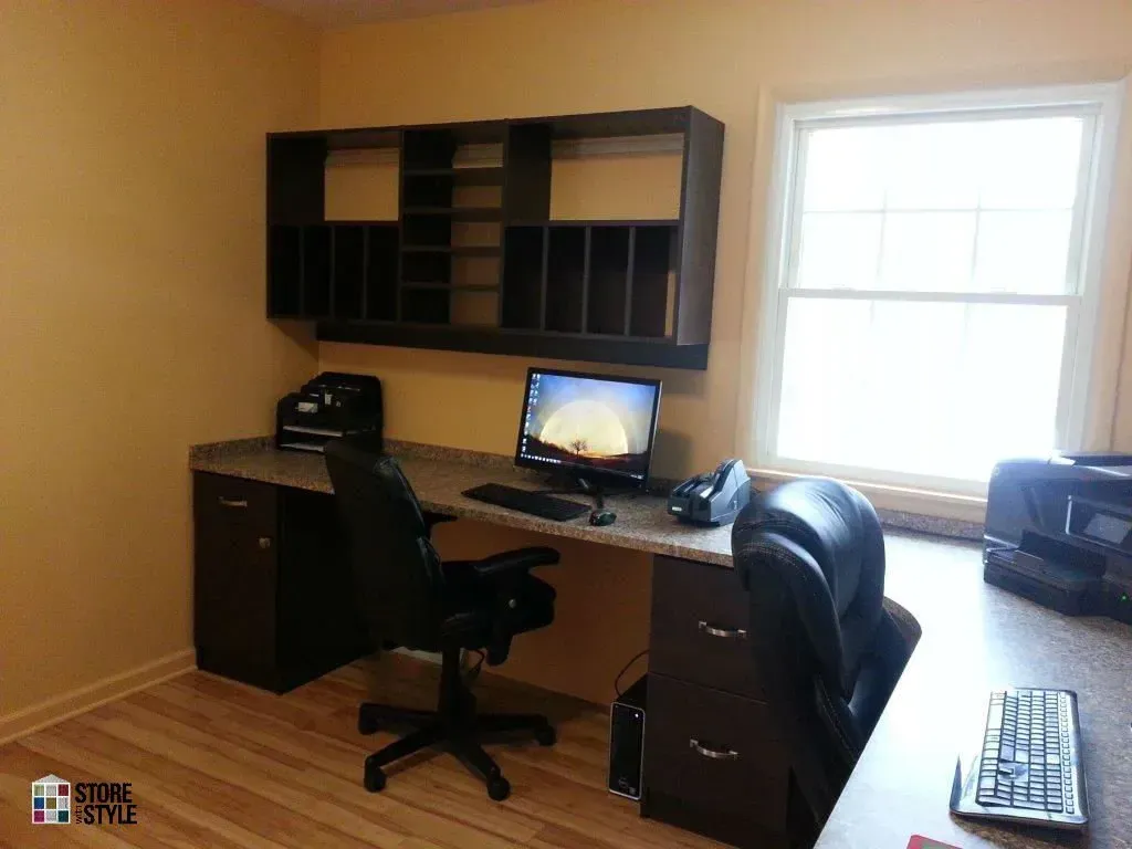 Office desk with a computer, chair, and wall-mounted storage, next to a window.