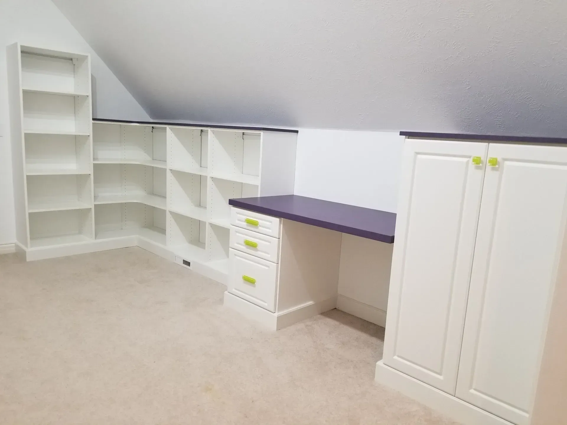 Built-in white shelving and desk in an attic space, with a purple desktop and green drawer pulls.