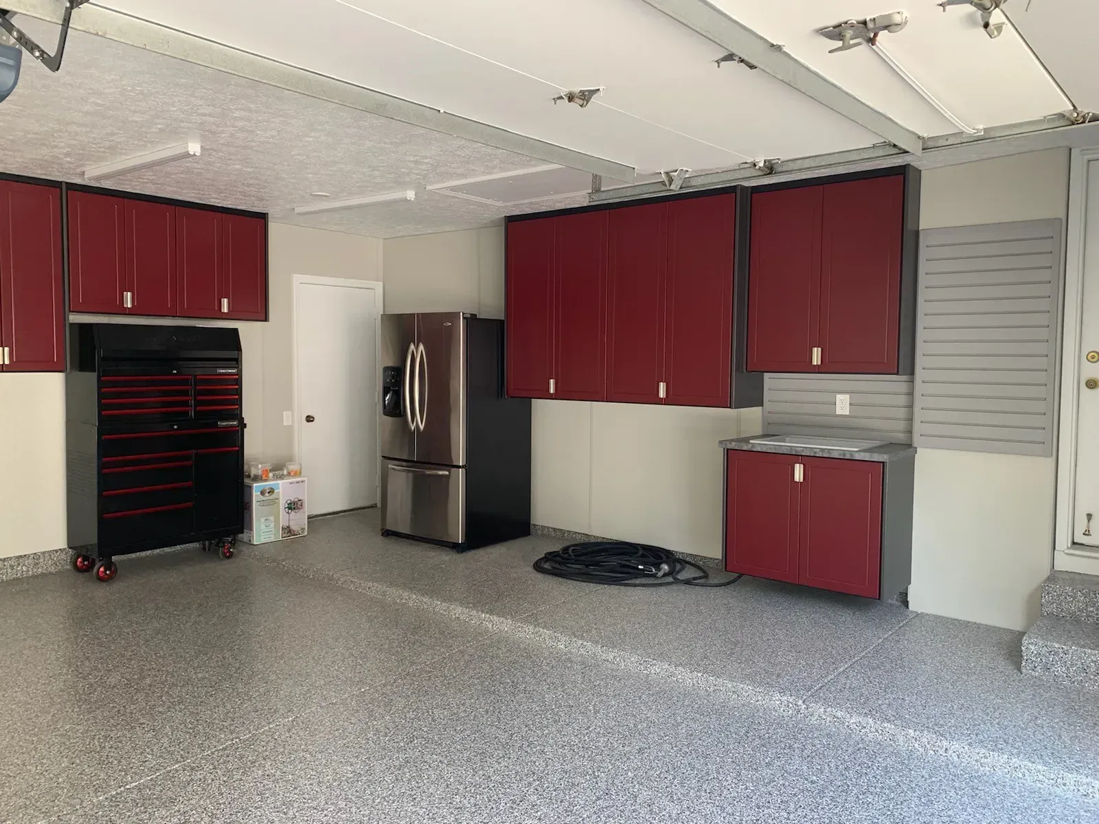 Garage with red cabinets, black toolbox, stainless steel fridge, gray flooring.
