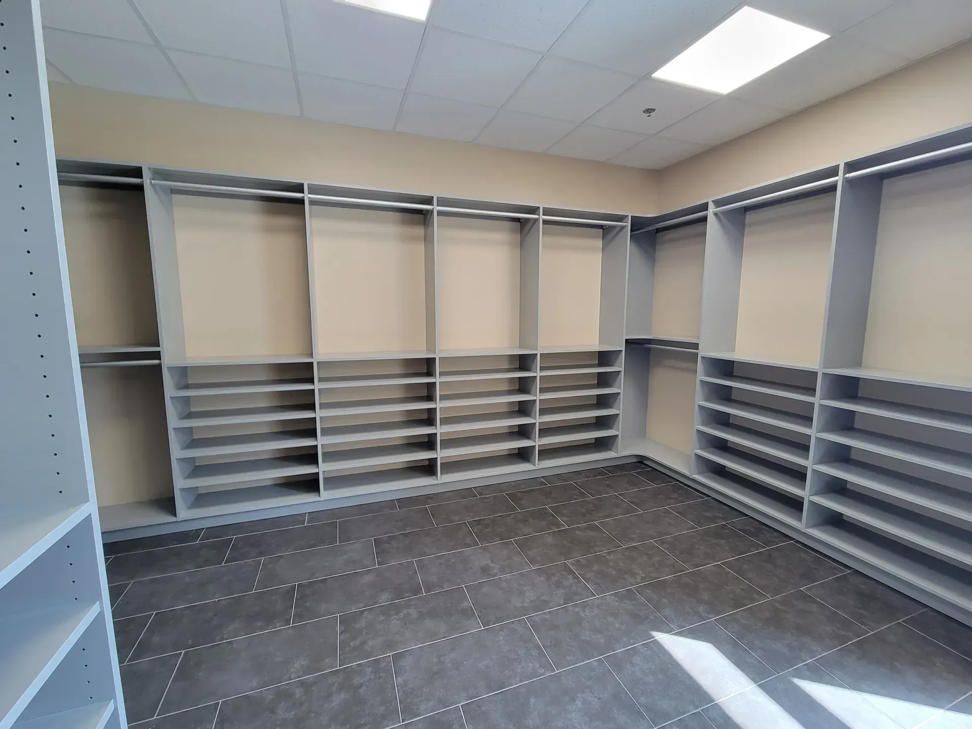 Empty walk-in closet with gray shelving and hanging rods, beige walls, and gray tiled floor.