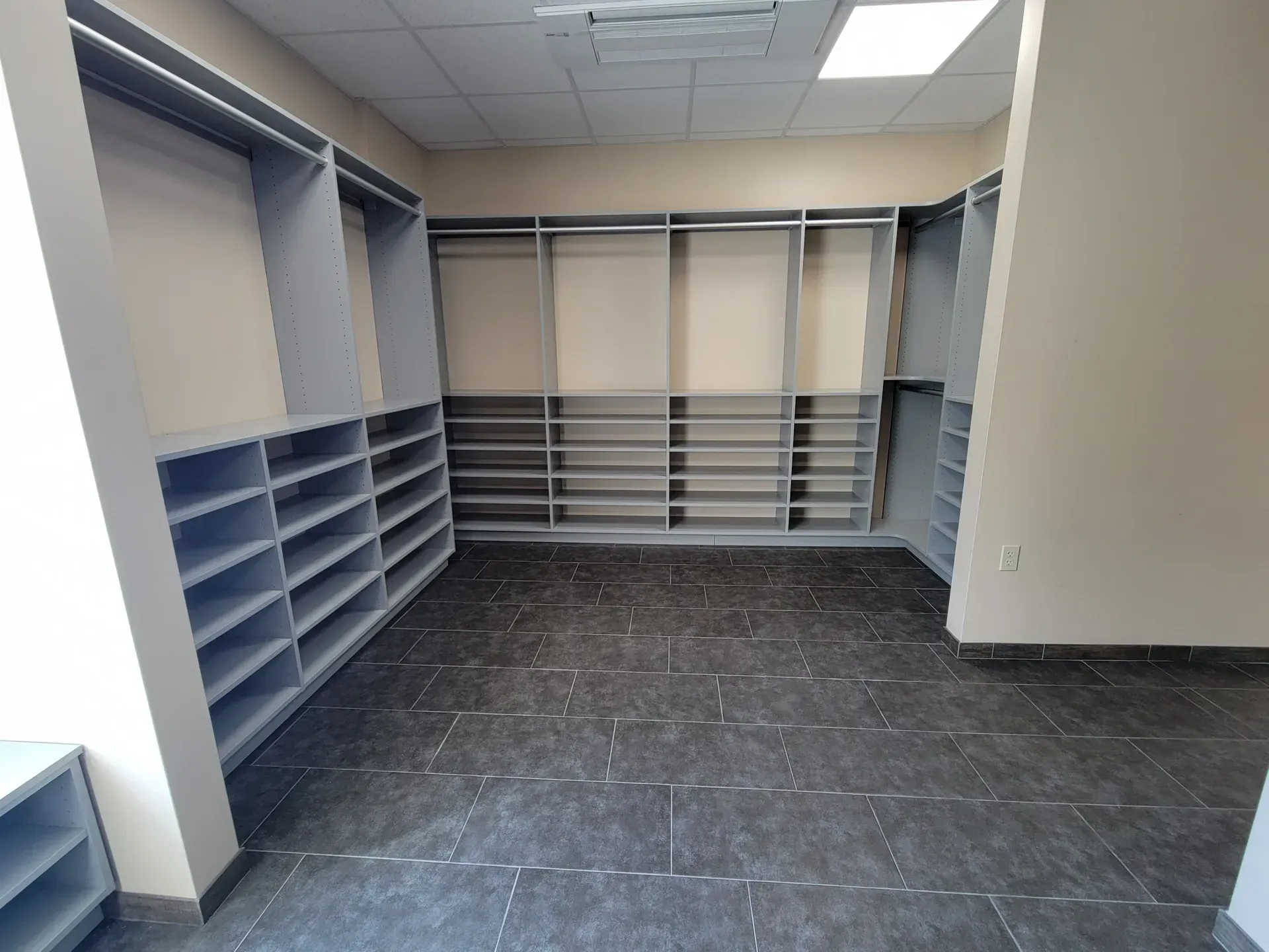 Empty walk-in closet with gray shelving units, beige walls, and dark tile floor.