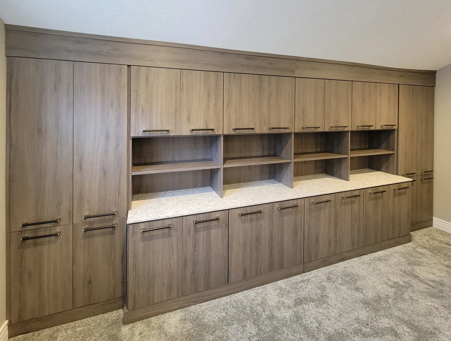 Built-in cabinetry with wood-look doors and open shelving, against a beige wall, in a room with carpet flooring.