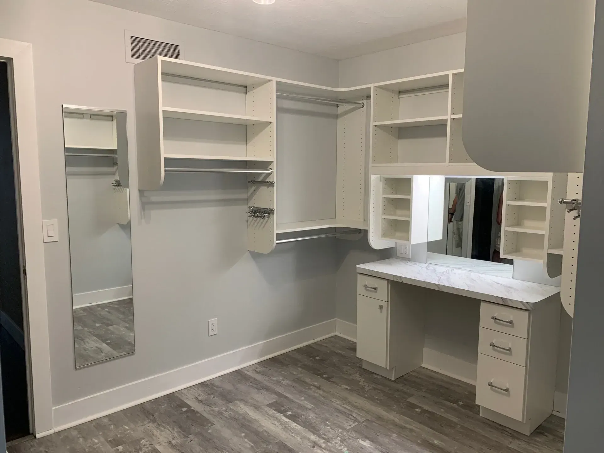 White custom closet with shelves, hanging rods, and a desk, in a room with gray walls and wood-look floor.