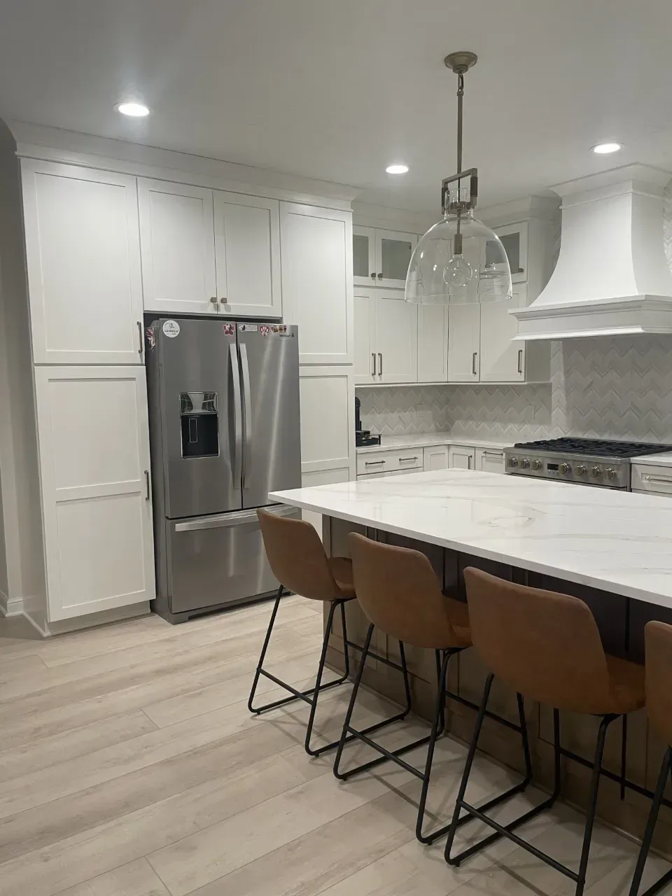 White kitchen with stainless steel refrigerator, island with brown bar stools, and pendant light.