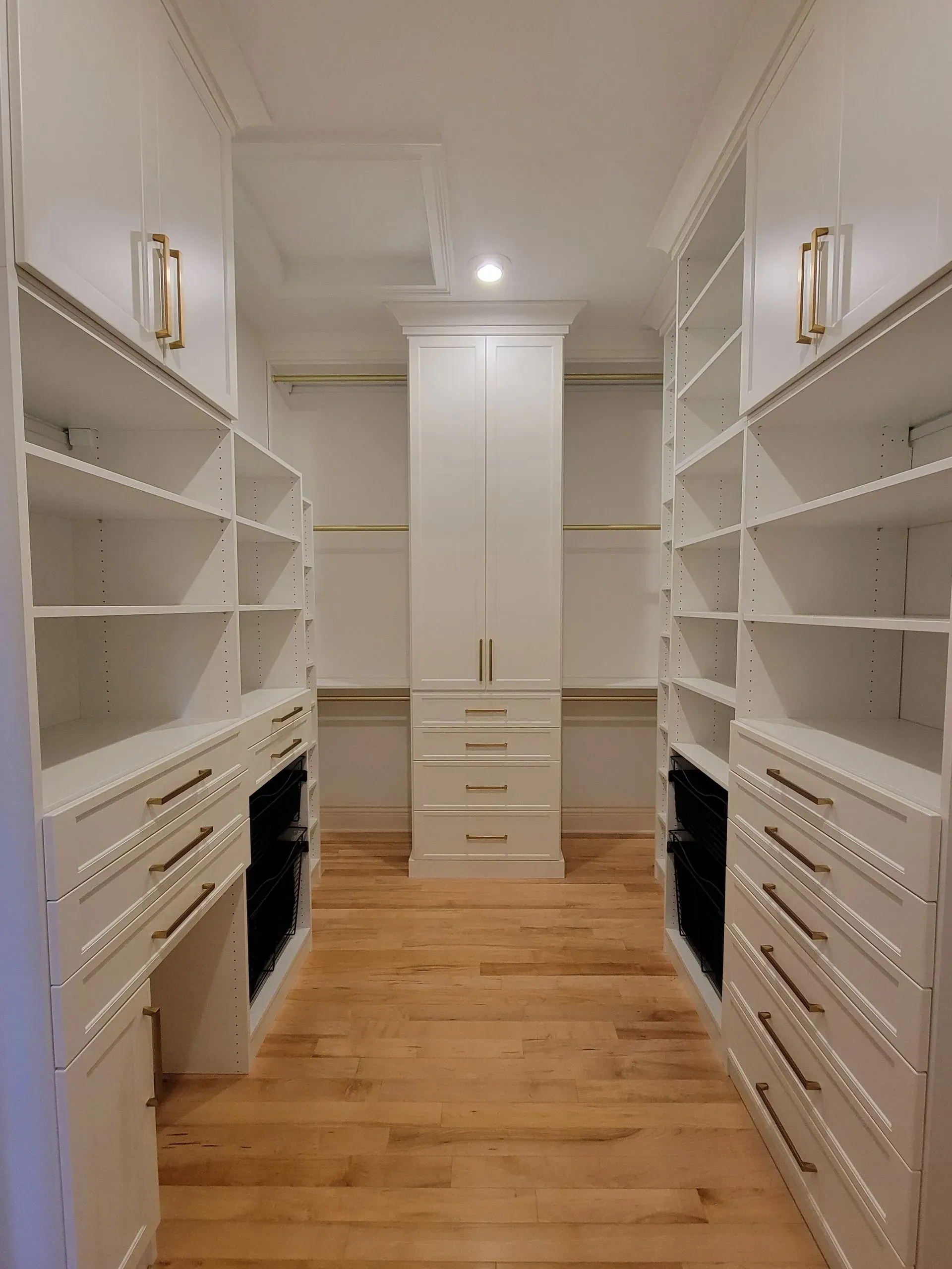 White walk-in closet with shelves, drawers, a central cabinet, and hardwood floors; golden light fixtures on the walls.