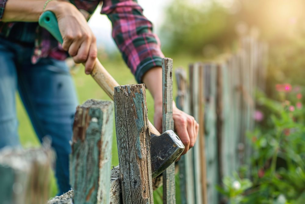 A person in a plaid shirt uses an axe to repair a weathered wooden fence in an outdoor setting.