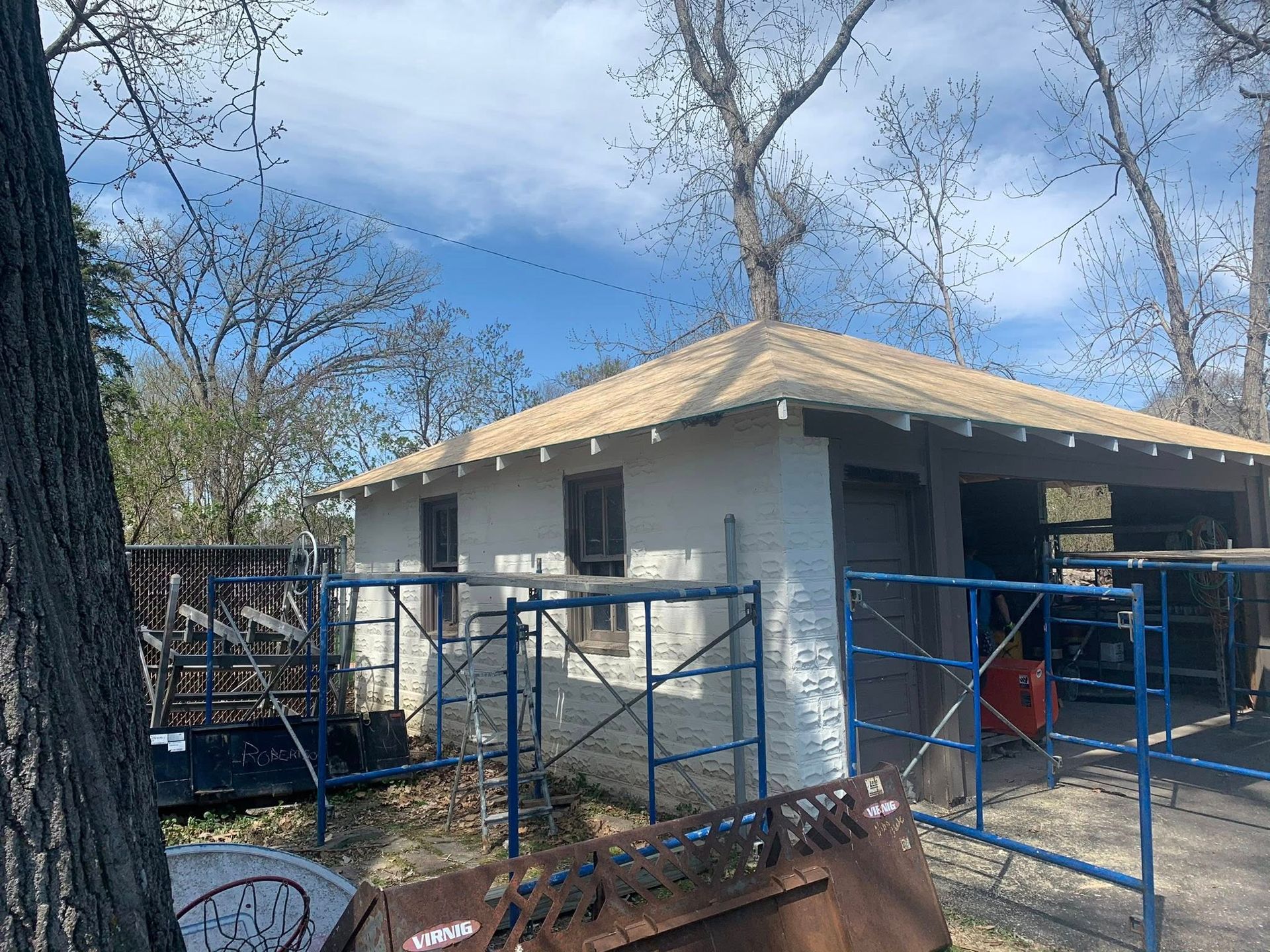 A small white building with a new shingled roof under construction, surrounded by blue scaffolding in a wooded area.
