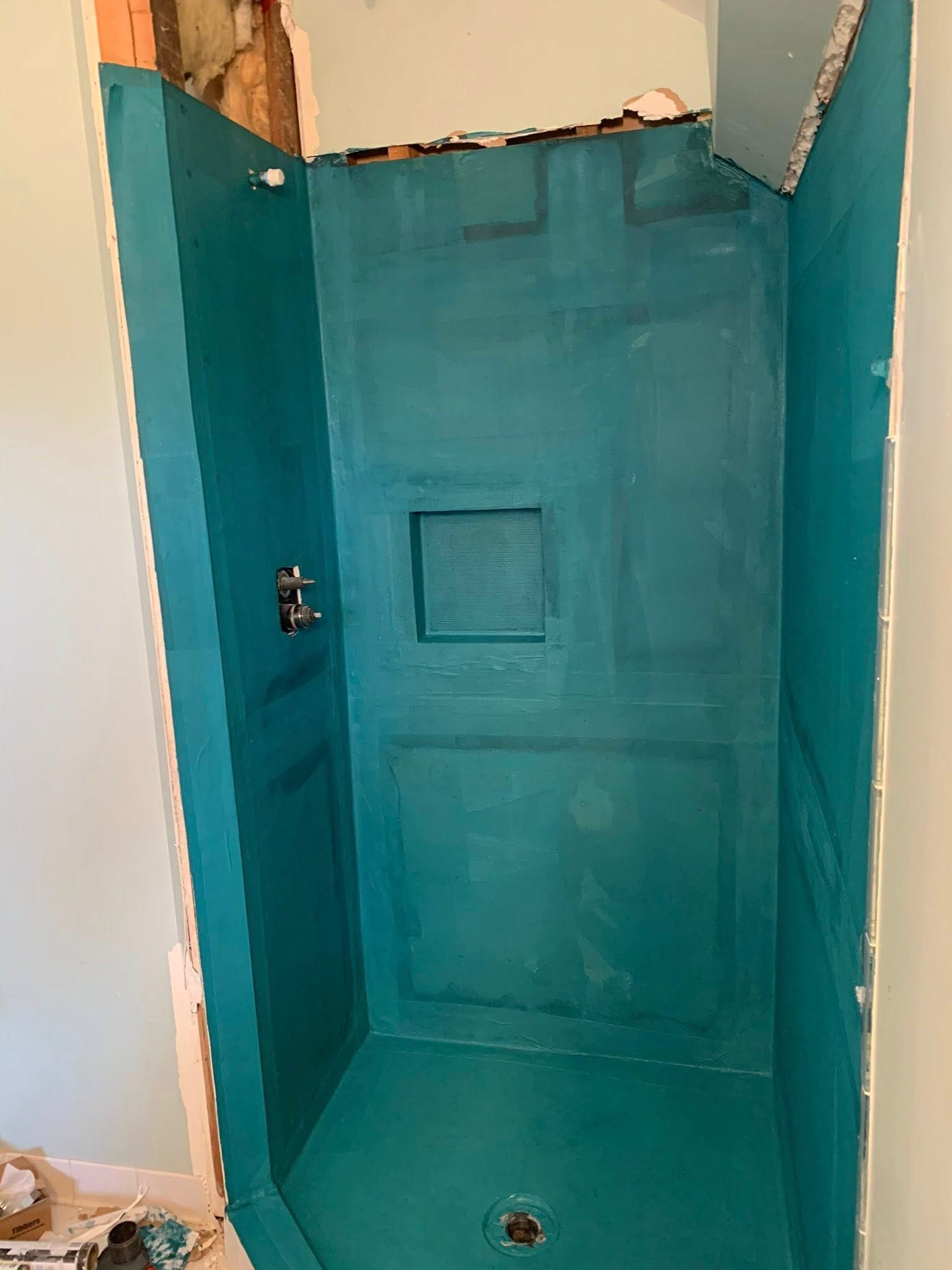 A teal-colored, waterproofed shower stall with a built-in niche and plumbing fixtures installed, mid-renovation.