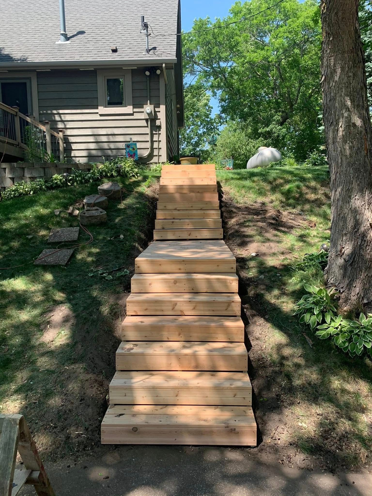 A straight, wooden staircase built into a grassy hillside leading up to the side of a grey house.
