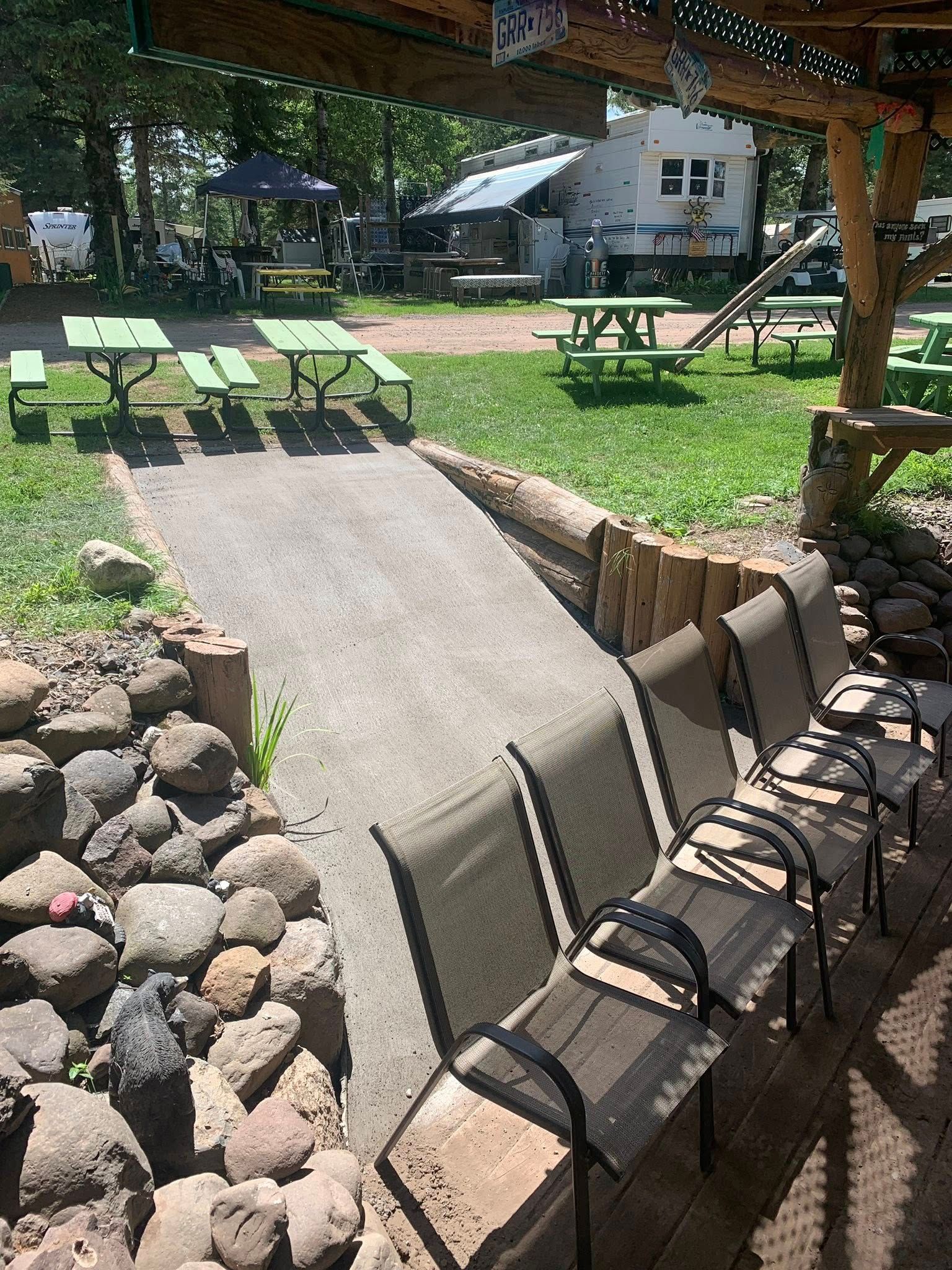 An outdoor seating area with rows of mesh chairs on a brick patio leading to a grassy space with picnic tables and an RV.