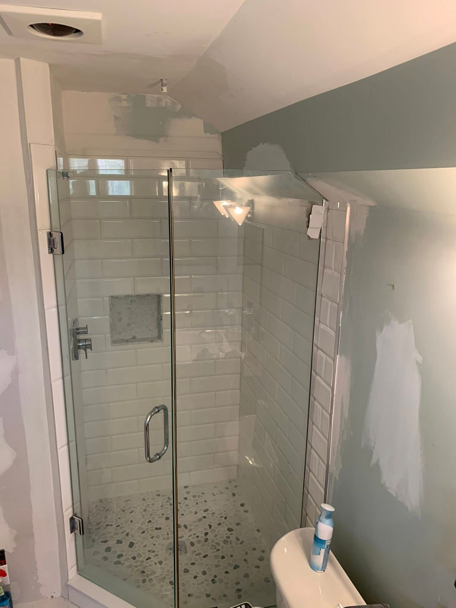 A corner glass shower stall with white subway tile, a recessed niche, and pebble tile flooring, in a bathroom under repair.