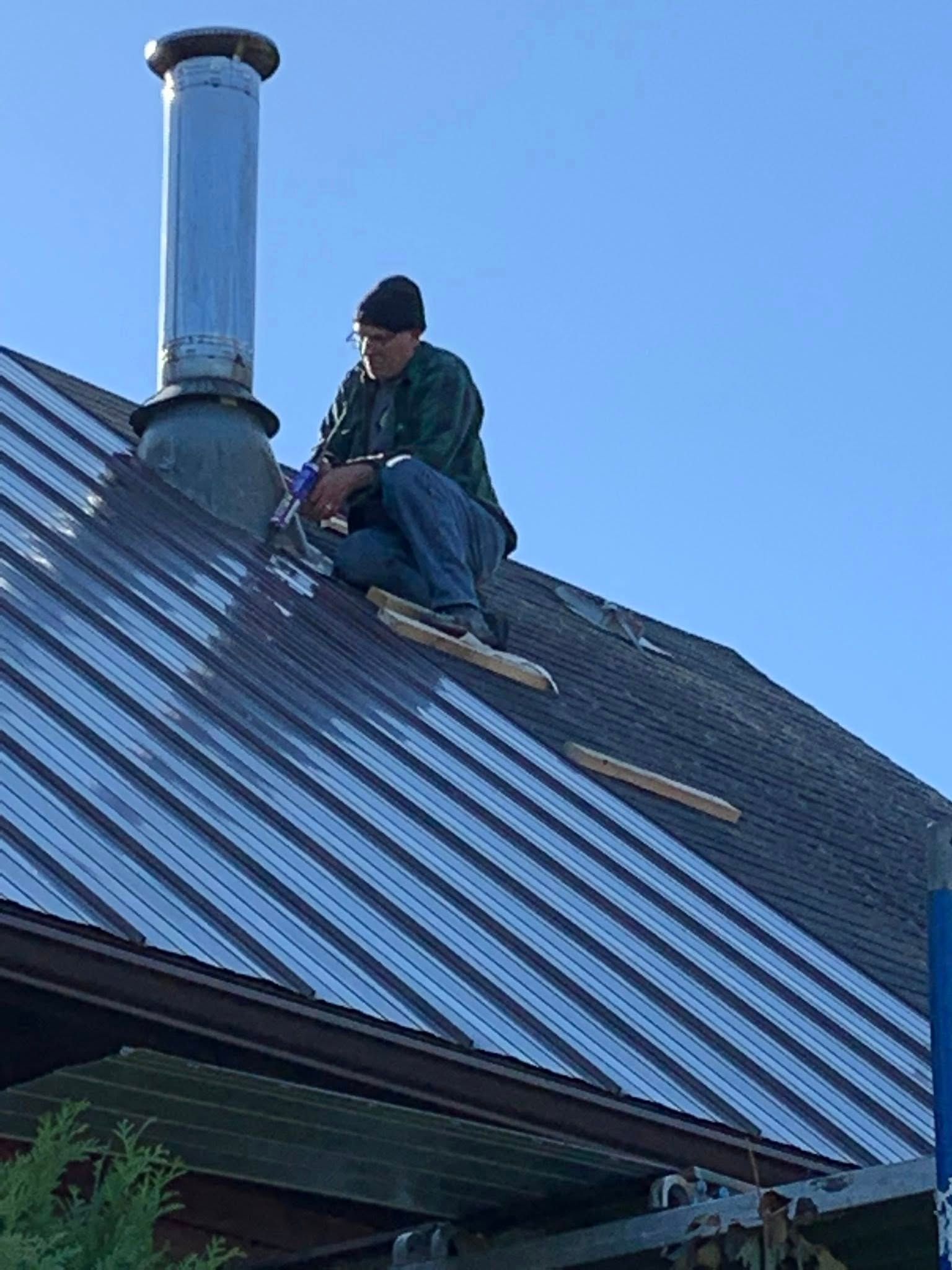 A person wearing a dark beanie and jacket works on a sloped metal roof next to a chimney under a clear blue sky.