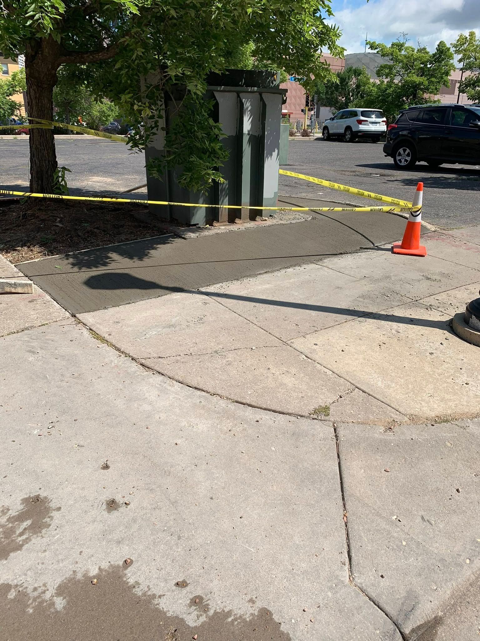 Yellow caution tape and an orange traffic cone surround a newly paved, dark patch of concrete by a utility box.