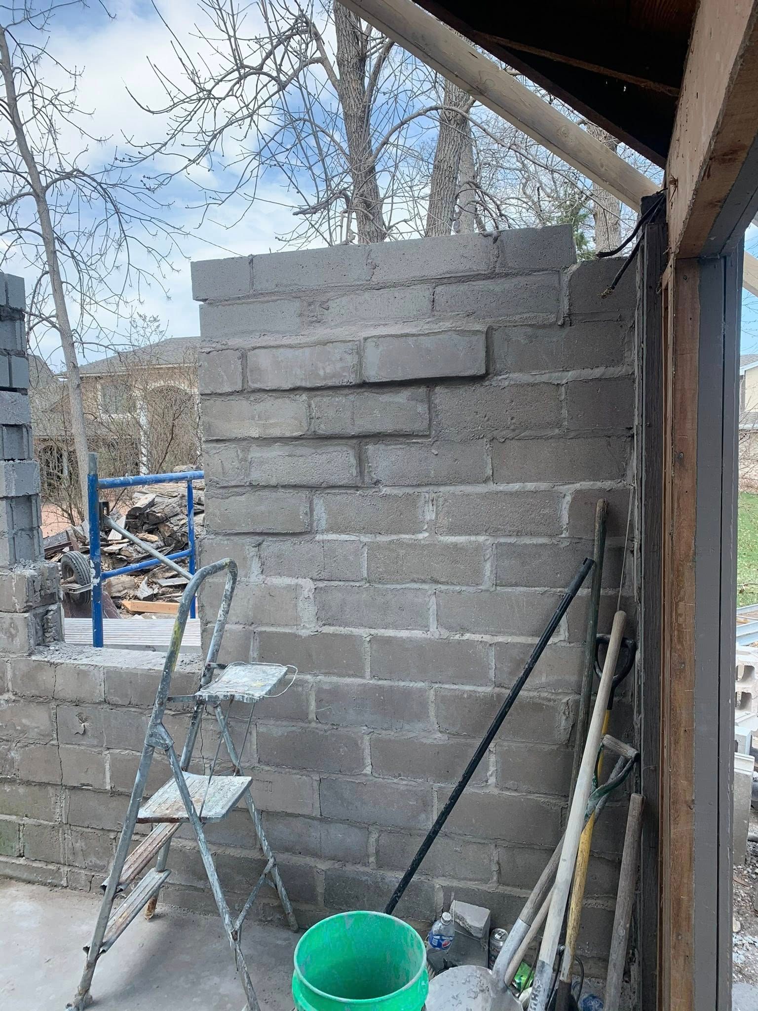 A partially constructed concrete block wall stands under a wooden roof frame, with a metal step ladder and bucket nearby.