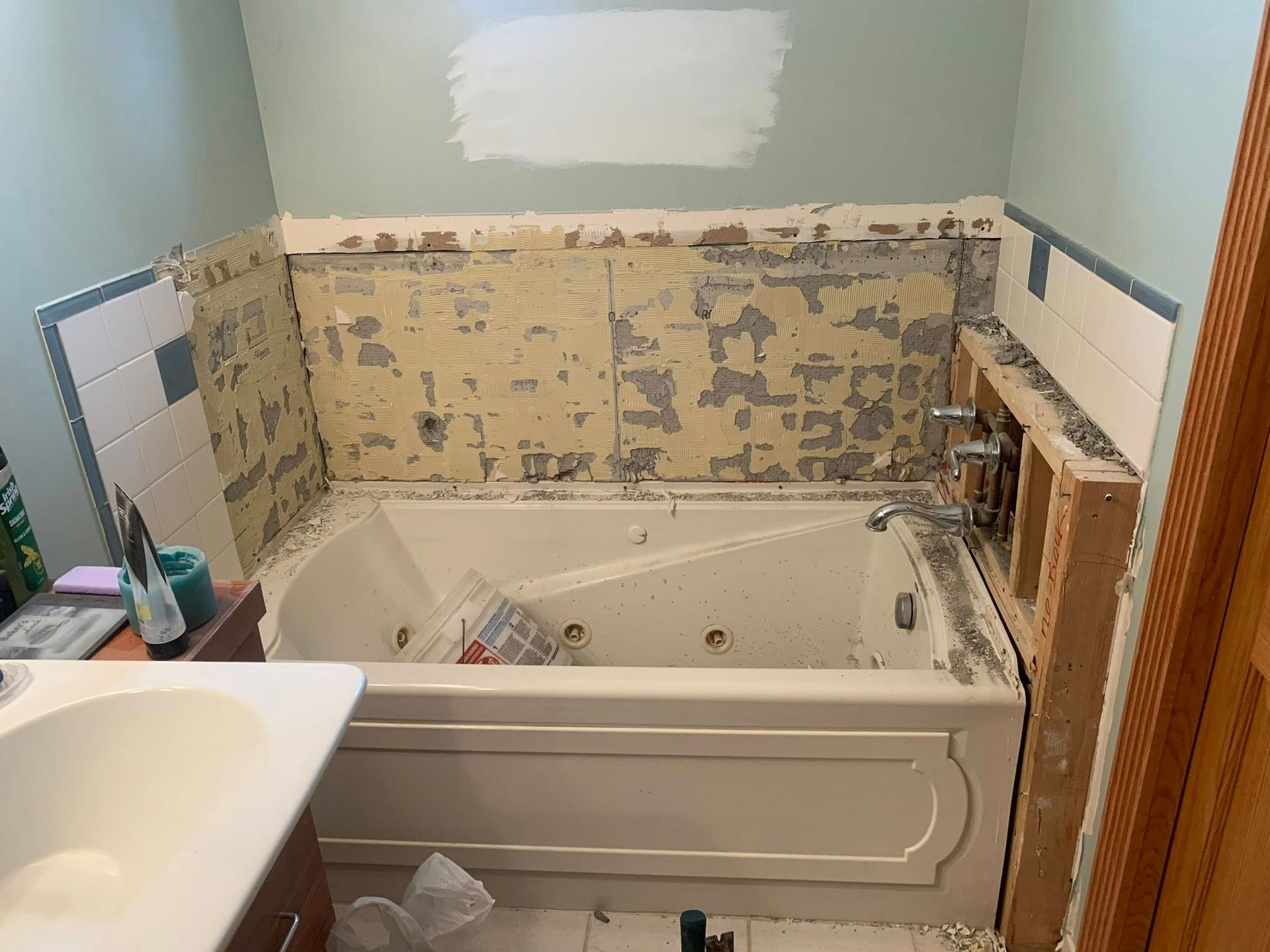 A bathroom under renovation featuring a jetted tub, exposed wooden framing near the faucets, and stripped wall tiles.