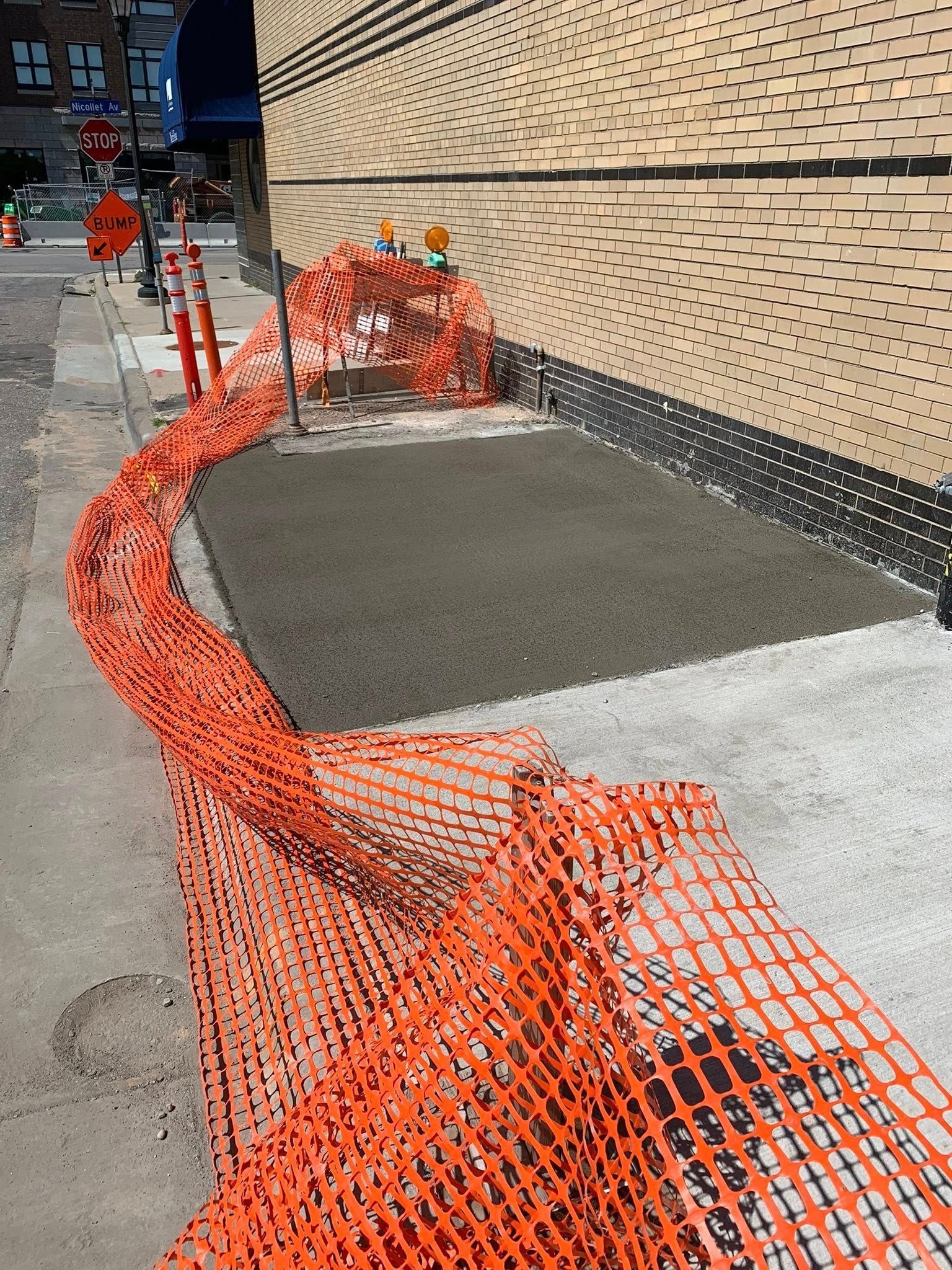 A freshly poured concrete patch on a sidewalk cordoned off by an orange plastic safety fence next to a building wall.