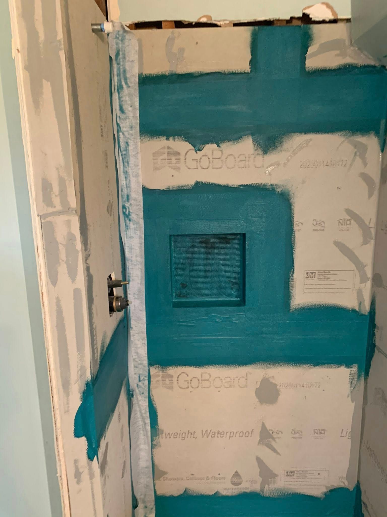 A partially tiled shower wall with turquoise sealant covering the seams of GoBoard panels and a recessed shower niche.