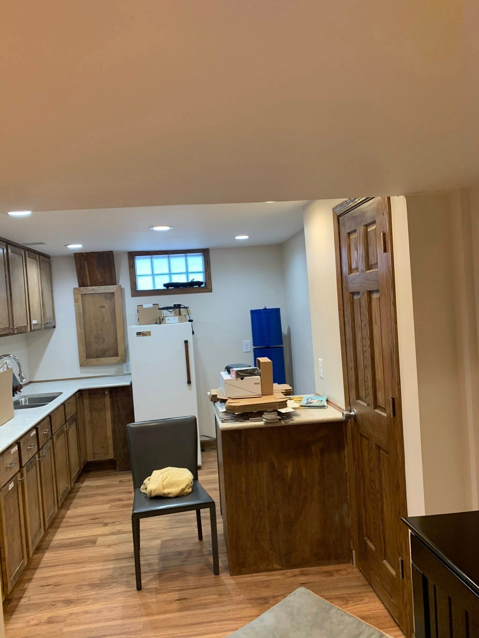 A kitchen with brown wooden cabinets, a white refrigerator, a gray chair, and a wooden door under a low ceiling.