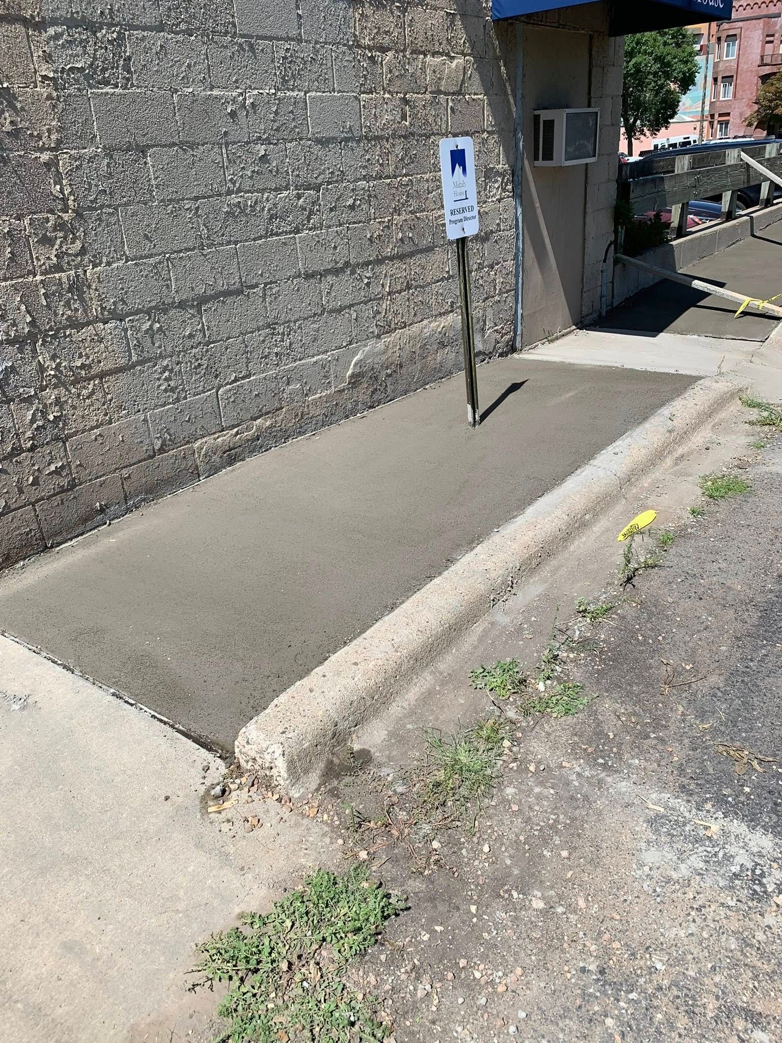 A handicap parking sign stands next to a freshly paved concrete patch against a grey stone wall near a building entrance.