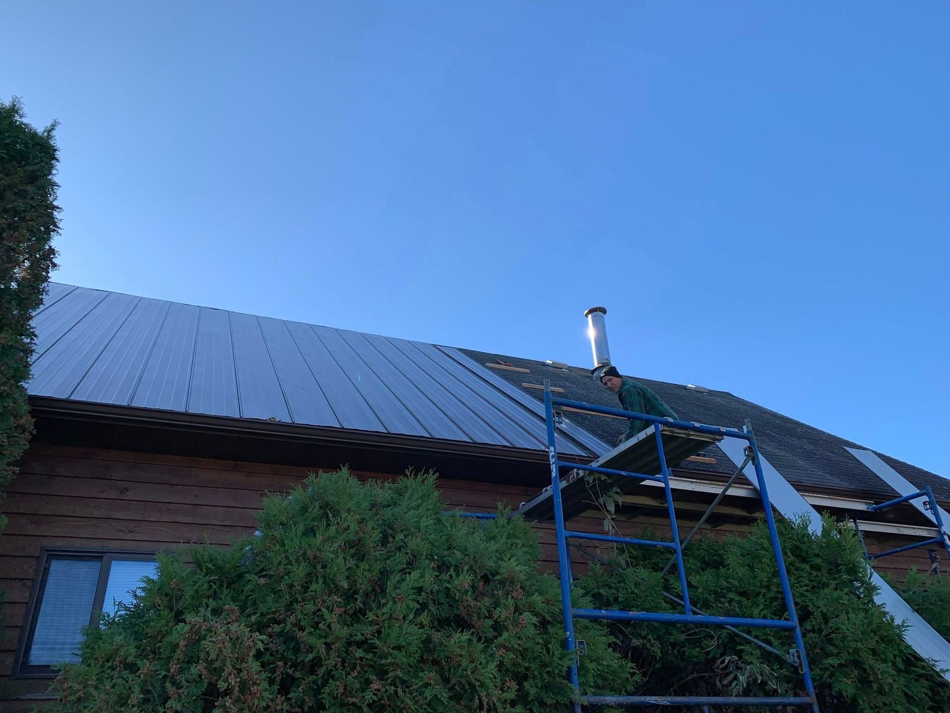 A worker on metal scaffolding repairs a house roof, installing metal panels beside an existing shingled section.