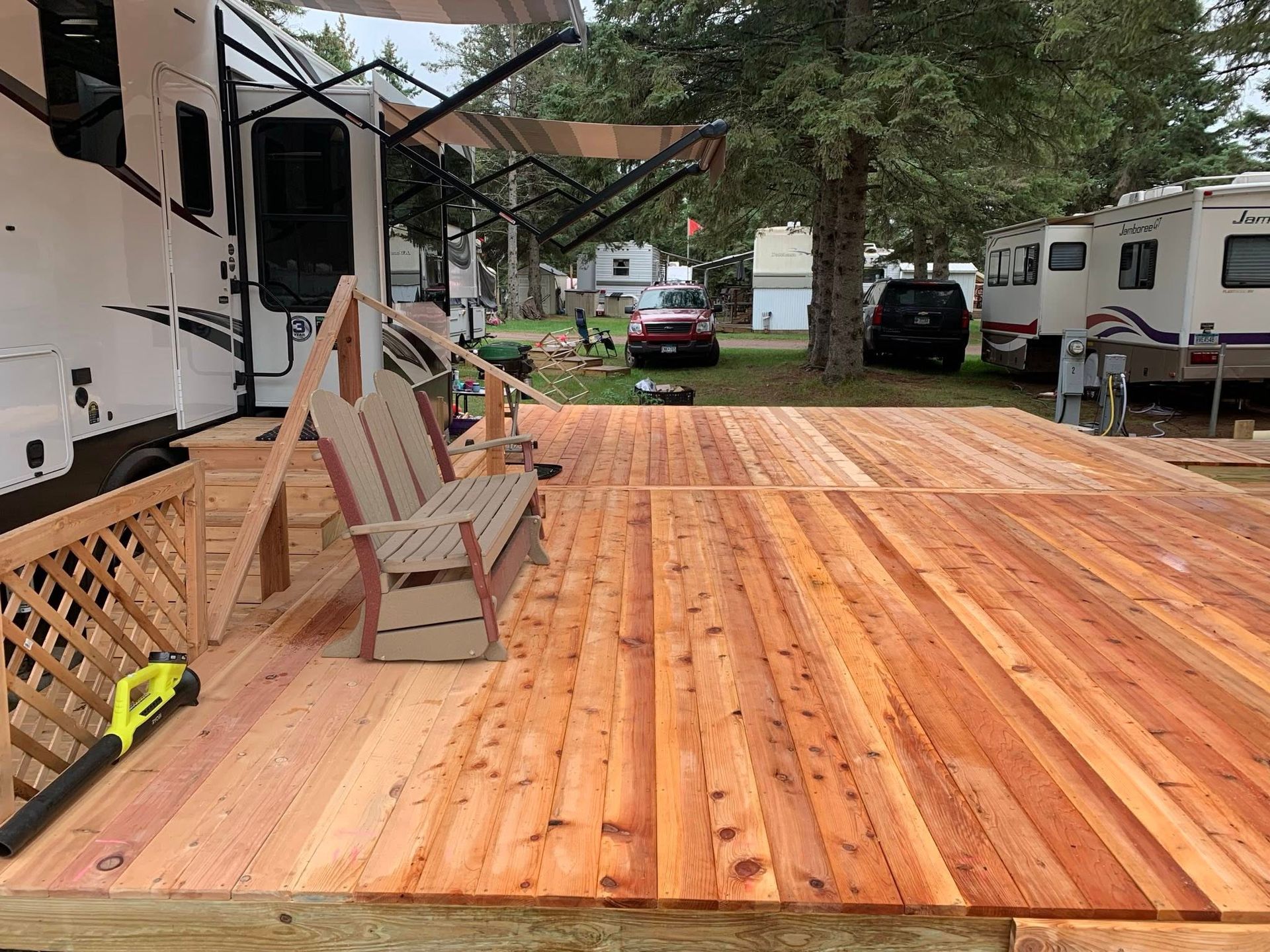 A wooden deck with a bench sits outside a large RV in a campsite, with other trailers and a car parked in the background.