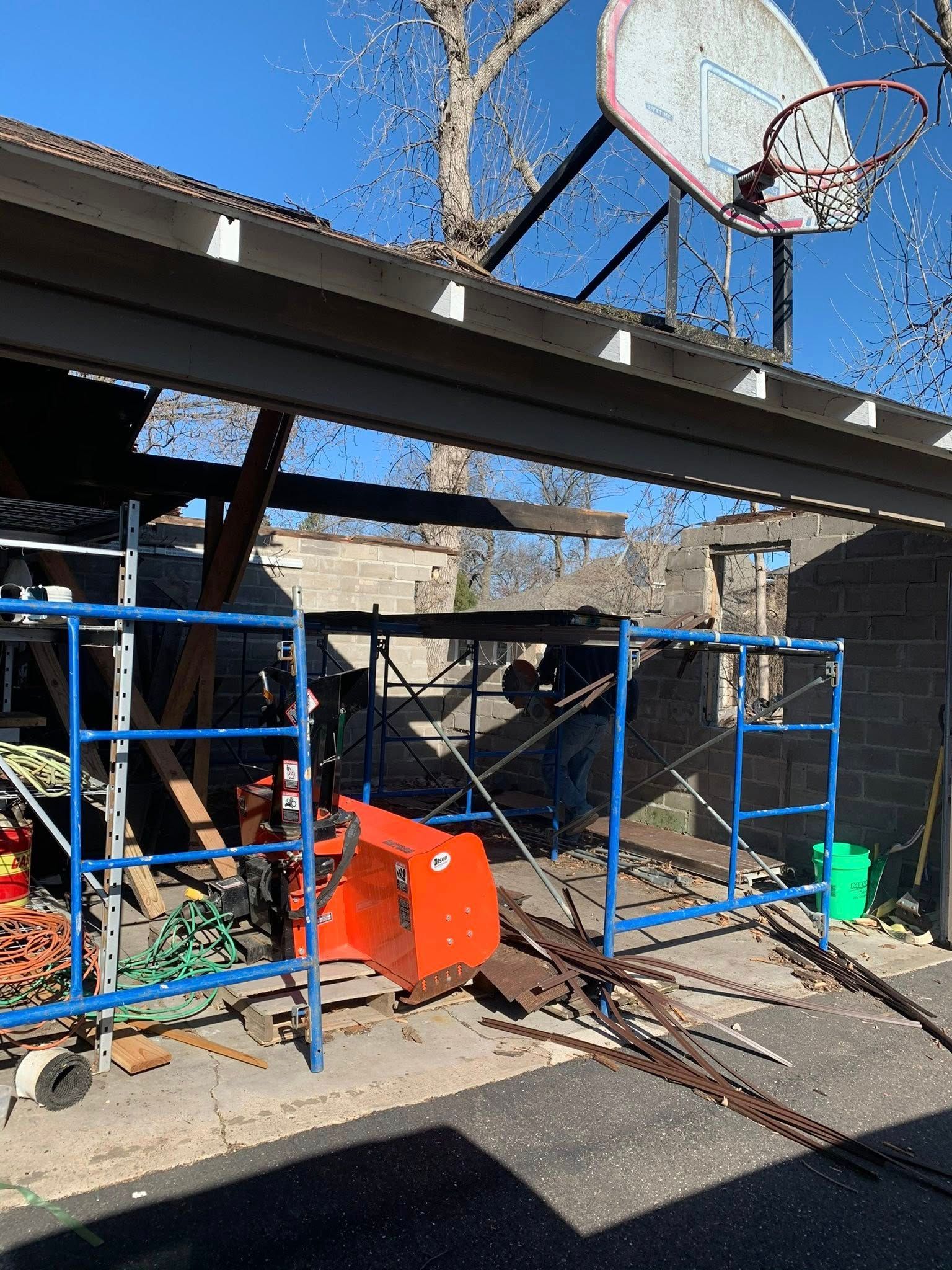 Construction scaffolding stands beneath a roof structure with a basketball hoop on a sunny day.