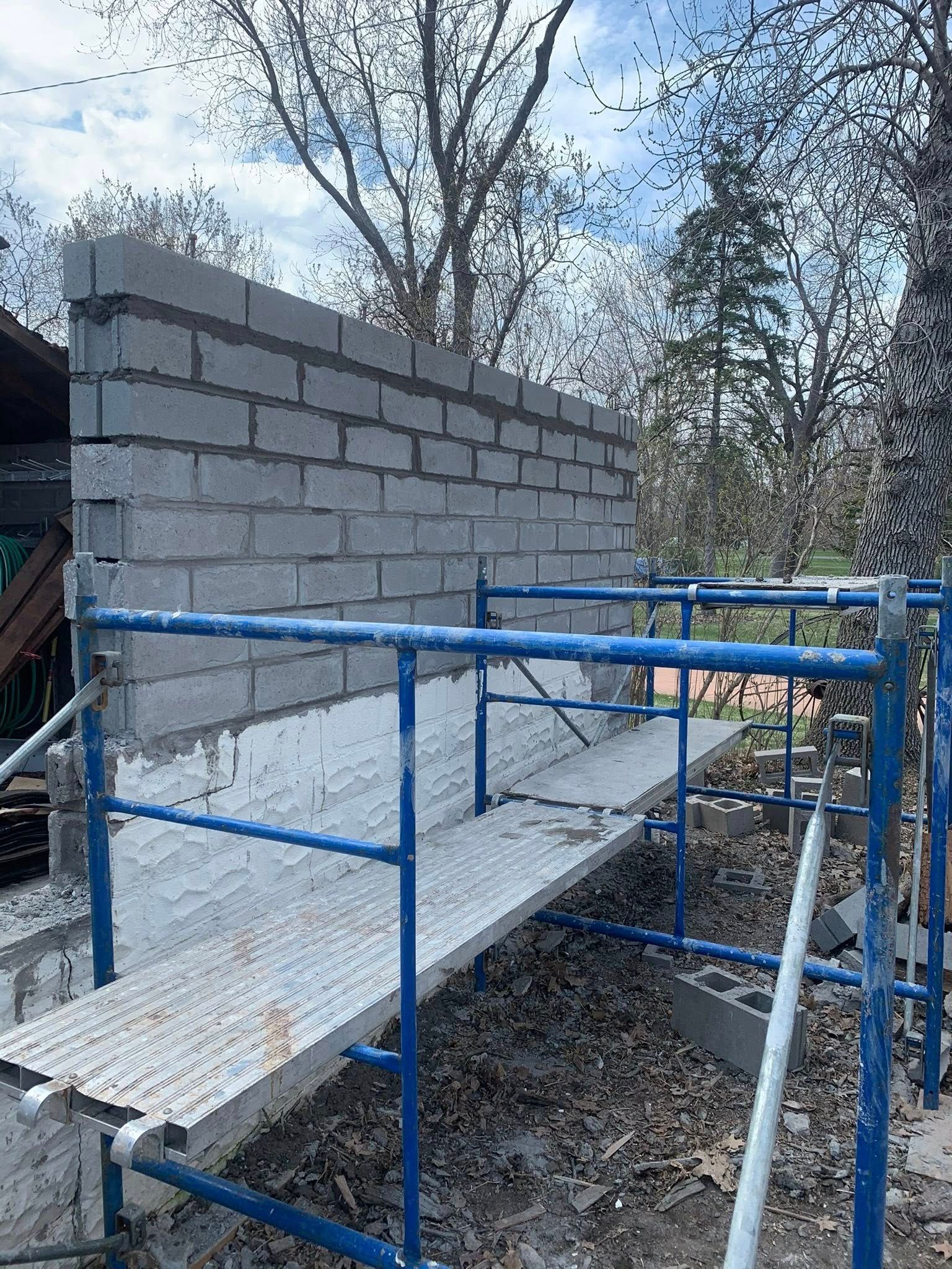 A partially constructed concrete block wall stands behind blue metal scaffolding on a gravel lot under a cloudy sky.