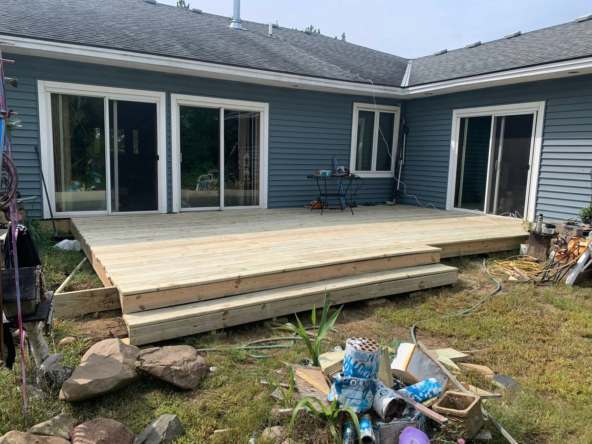 A newly constructed light-wood deck with two stairs attached to the back of a blue-sided house with three sliding doors.
