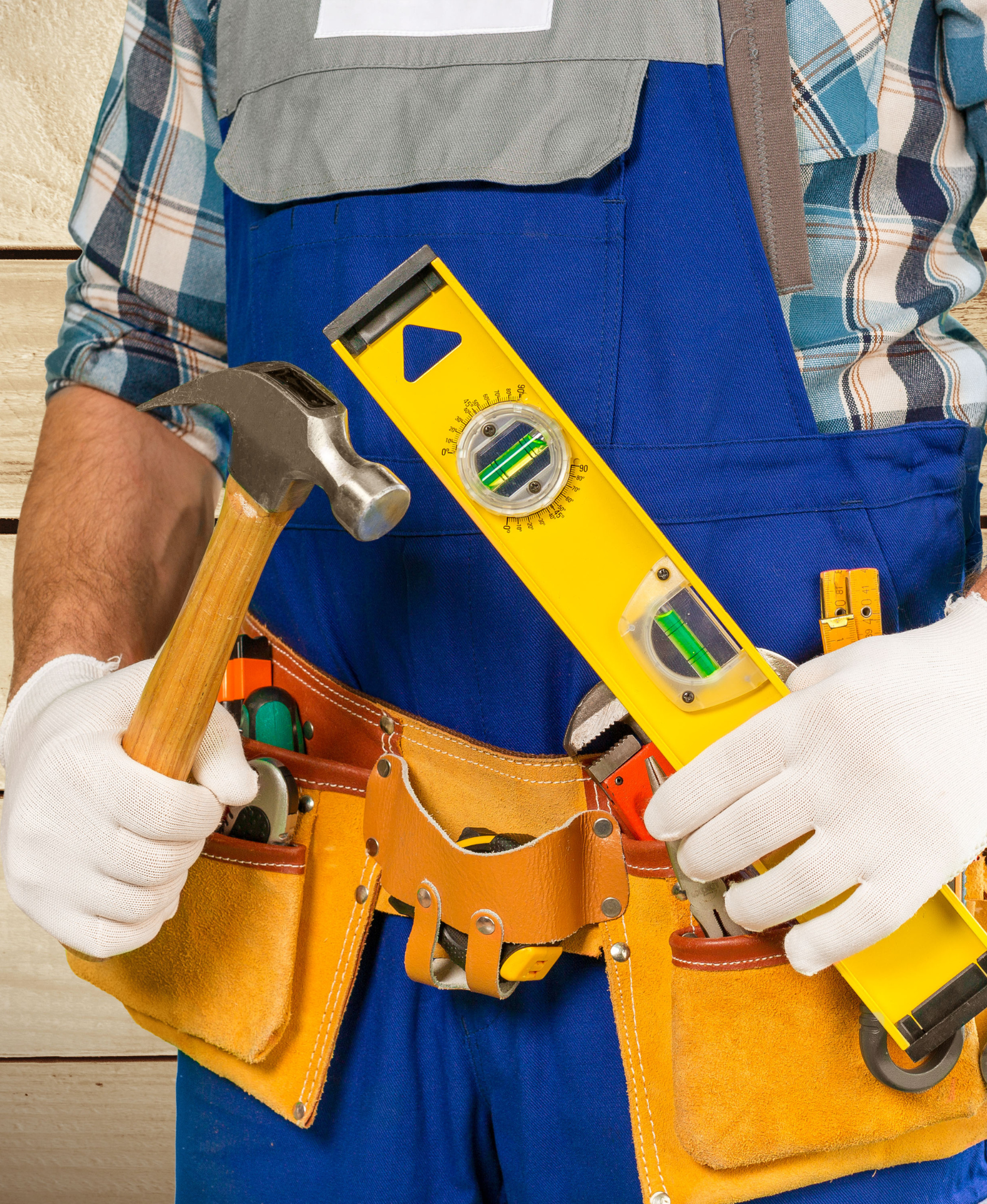 A person in blue coveralls and white work gloves holds a metal hammer and a yellow spirit level in a tool belt.