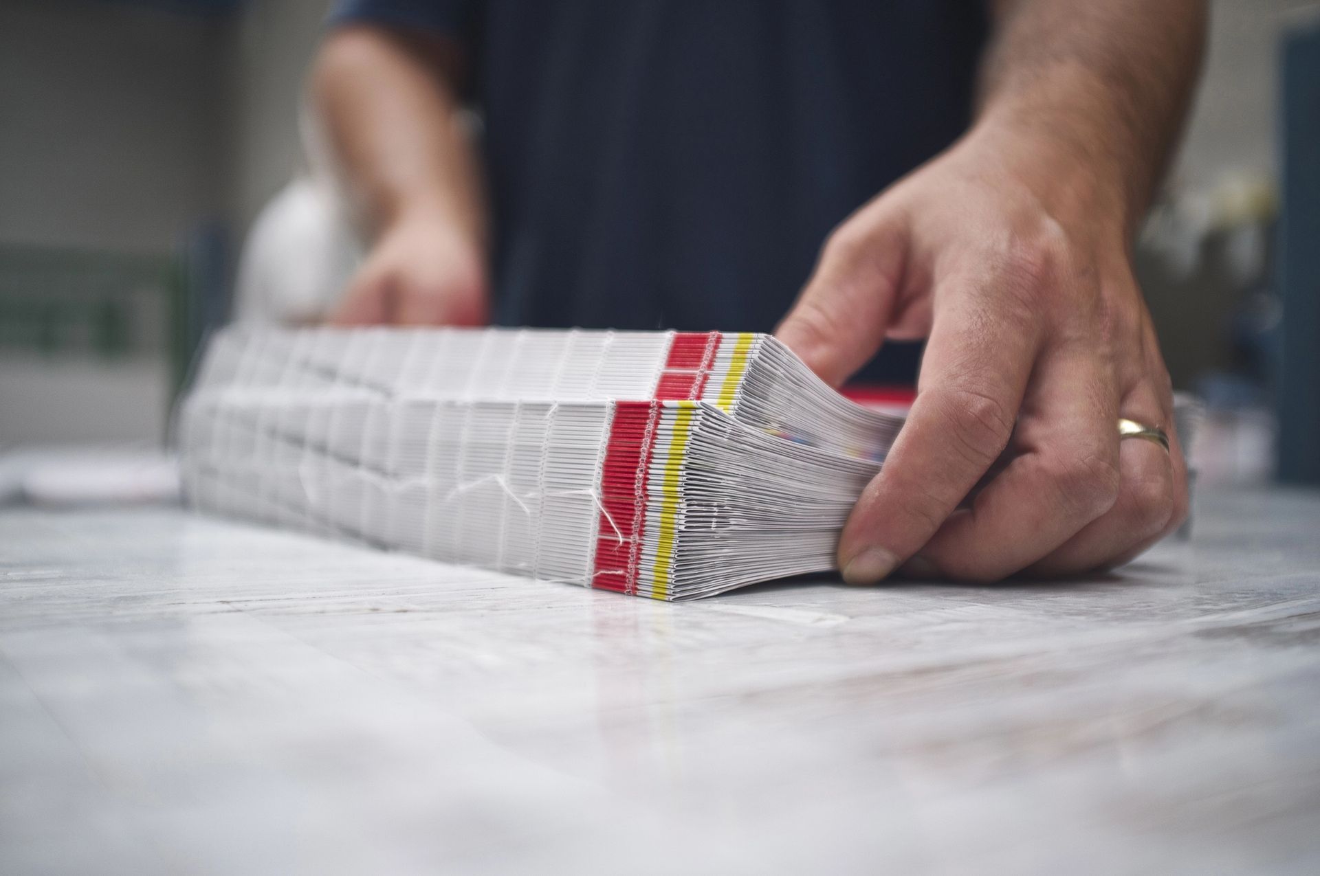 Mani di una persona che regge una pila di materiale stampato, strettamente legato con nastro rosso e giallo, su un tavolo.