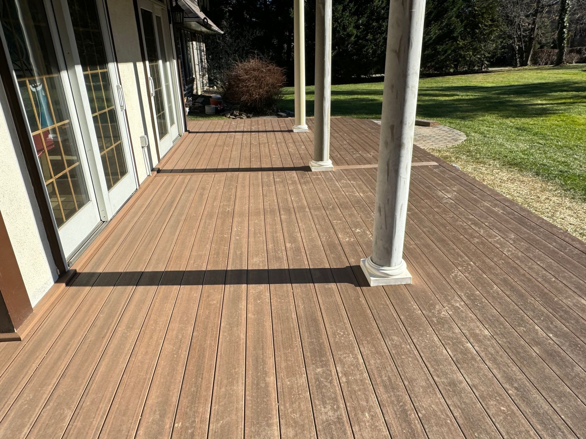 Brown composite deck with vertical grooves, under a covered porch with white columns, near a grassy lawn.