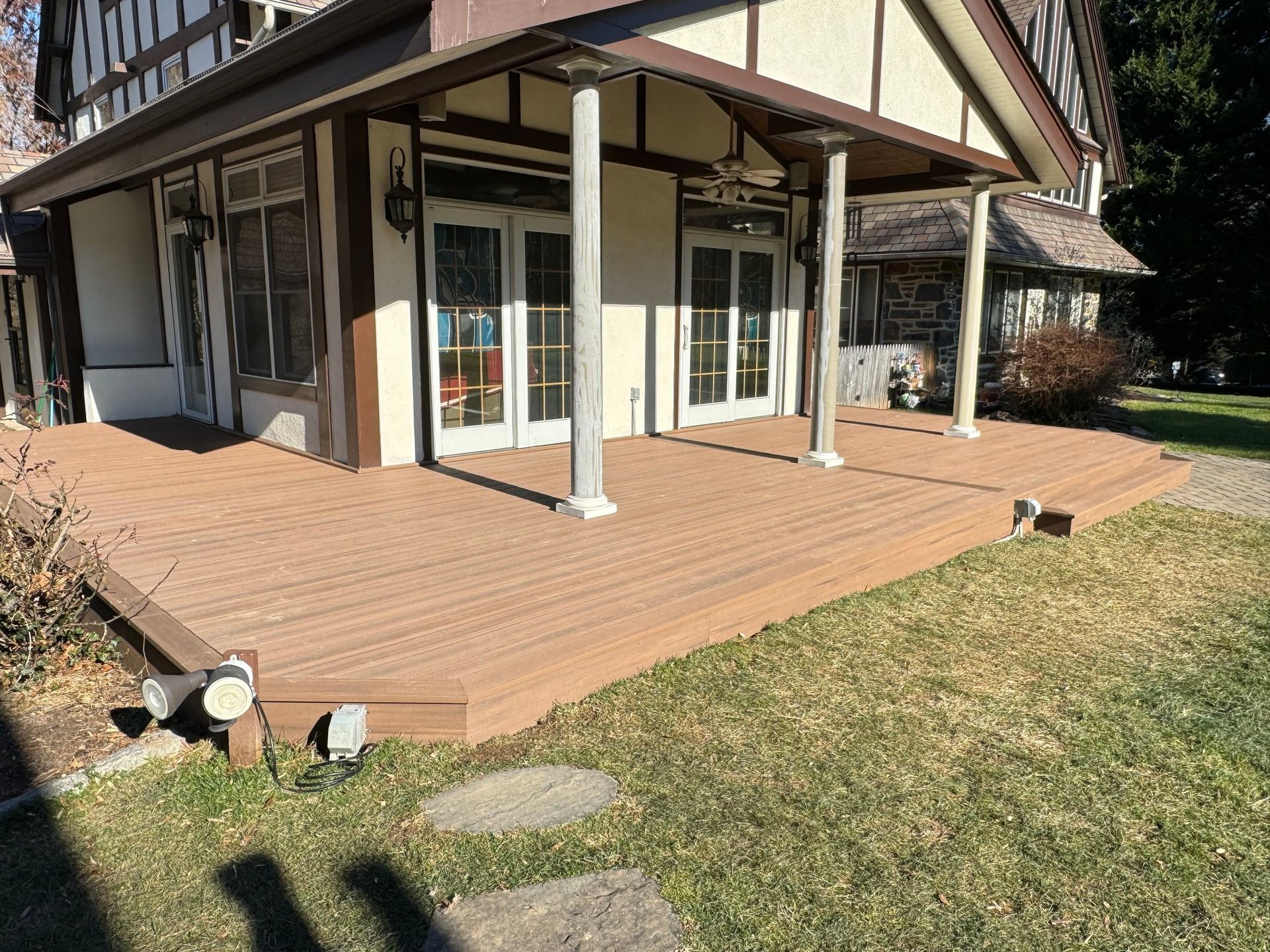 Brown wooden deck with a building featuring white columns and glass doors. Green grass and stone path in front.